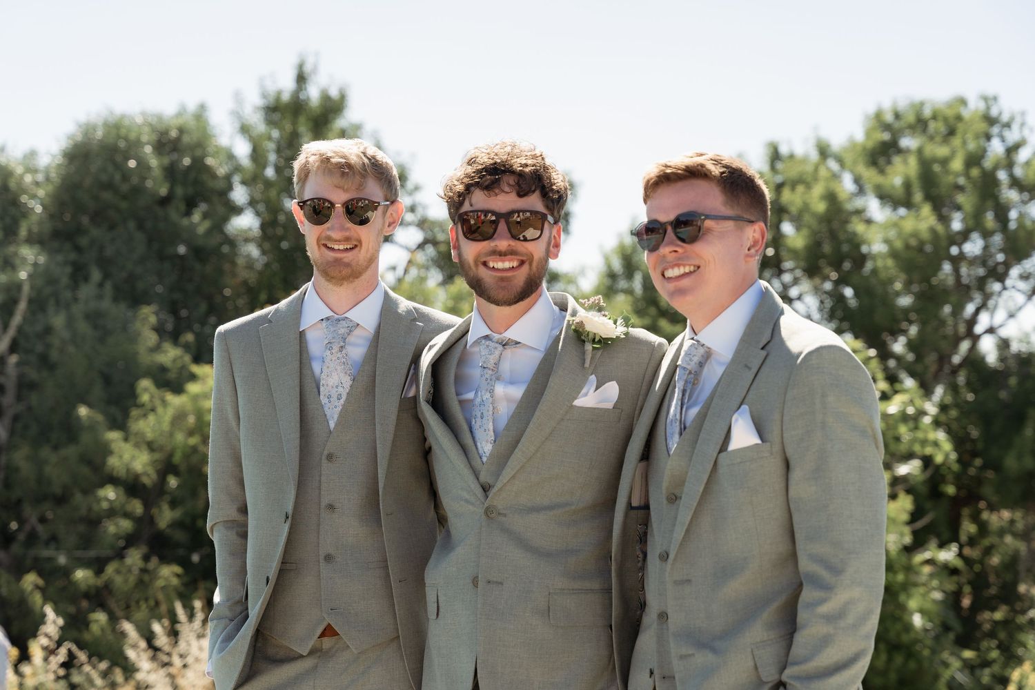 Three groomsmen in matching sage green suits and sunglasses smiling outdoors on a sunny wedding day.