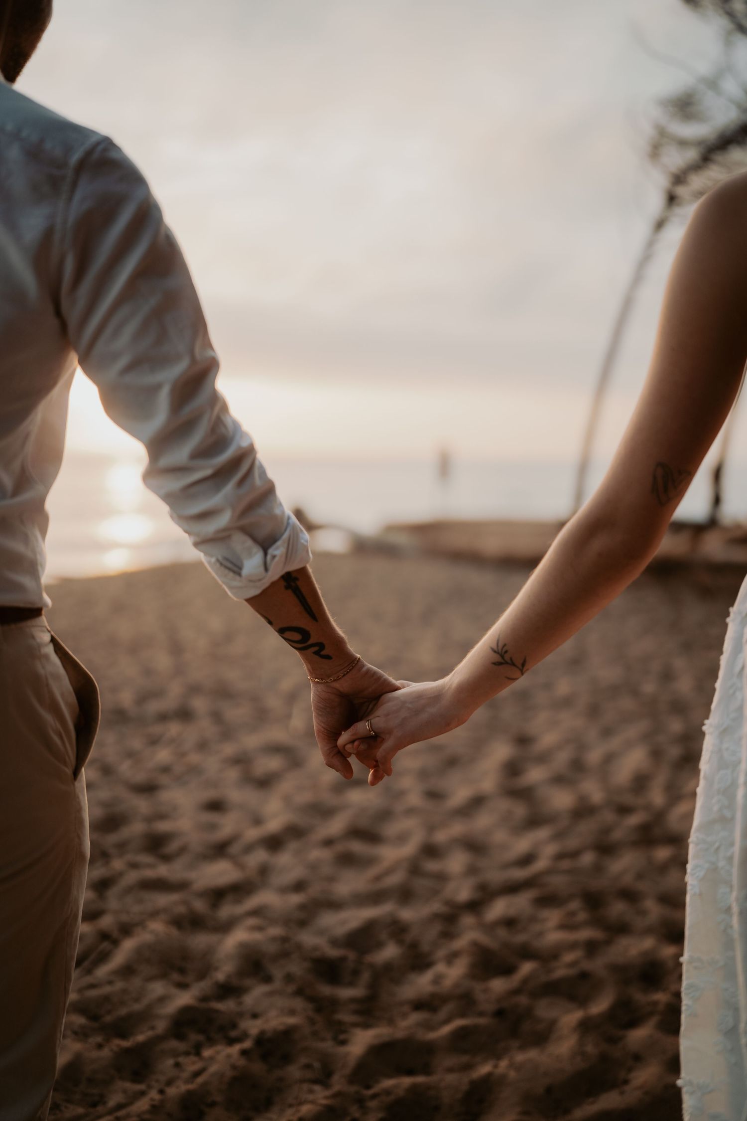 Couple holding hands on sandy beach at sunset, man with tattoos wearing white shirt and khaki pants.