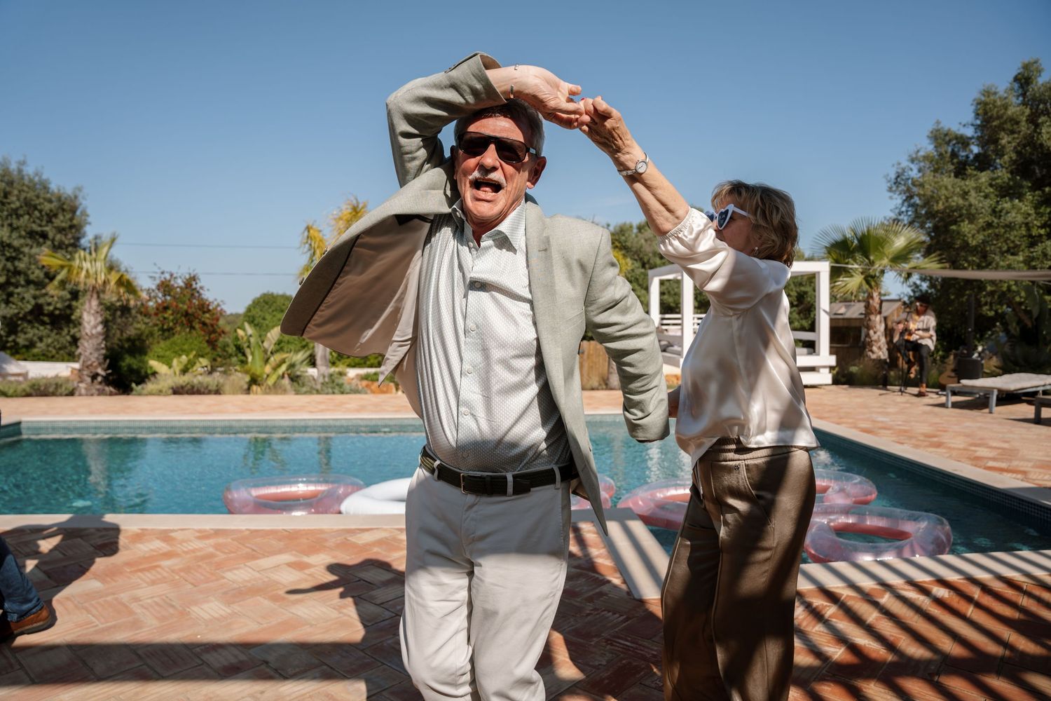 Two men dancing and celebrating outdoors by a pool at a sunny wedding reception.