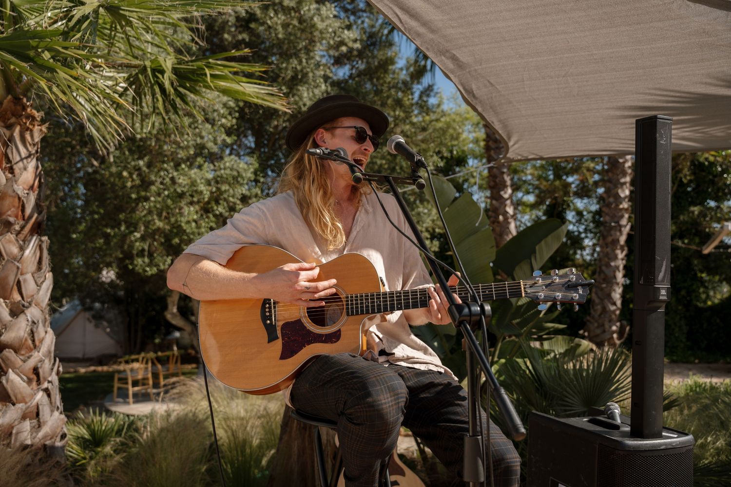 Musician in wide-brim hat playing acoustic guitar and singing at outdoor wedding under canopy.