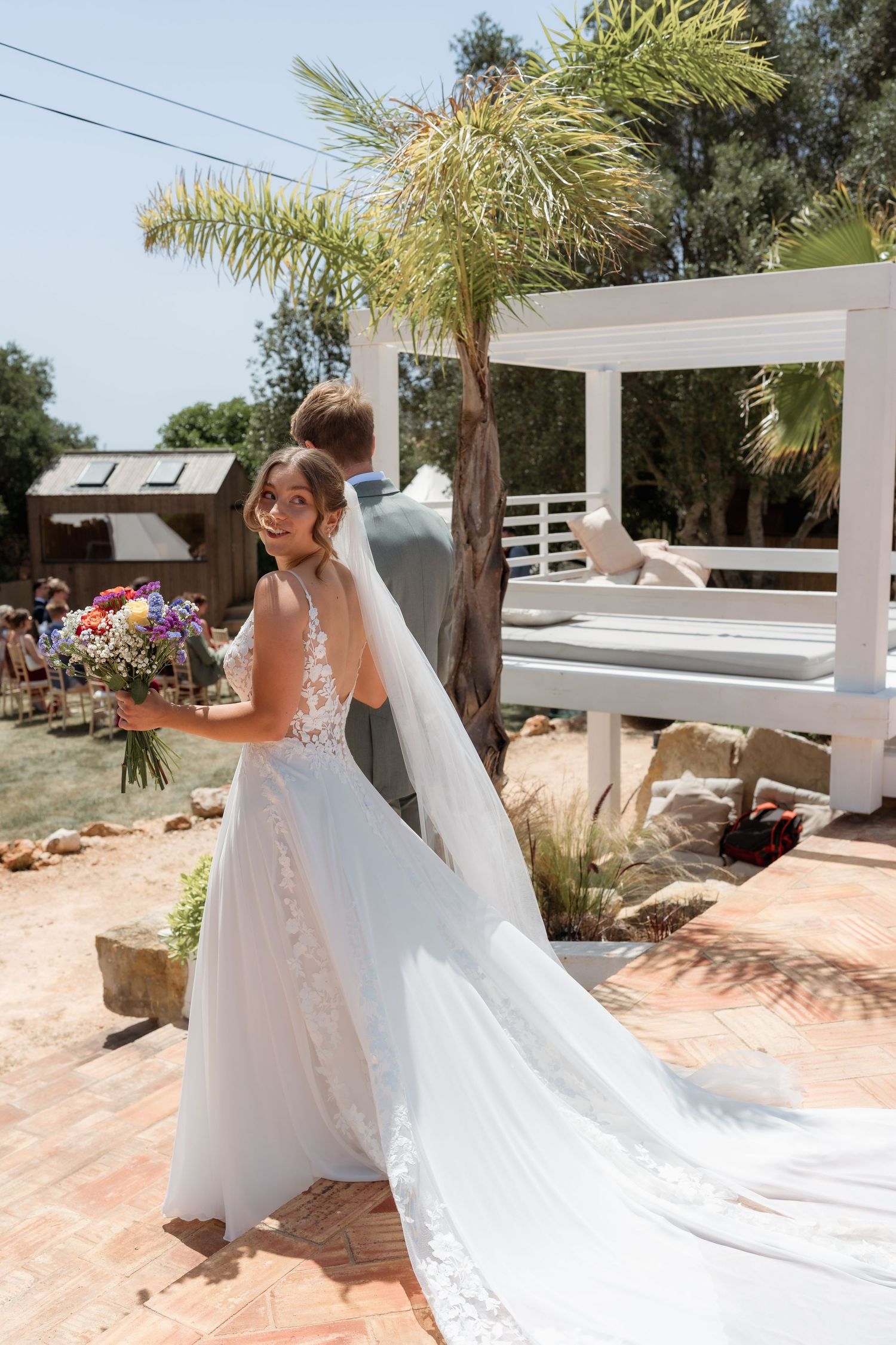 Bride in white gown with veil holds colorful bouquet, glancing back during outdoor wedding ceremony near palm tree.