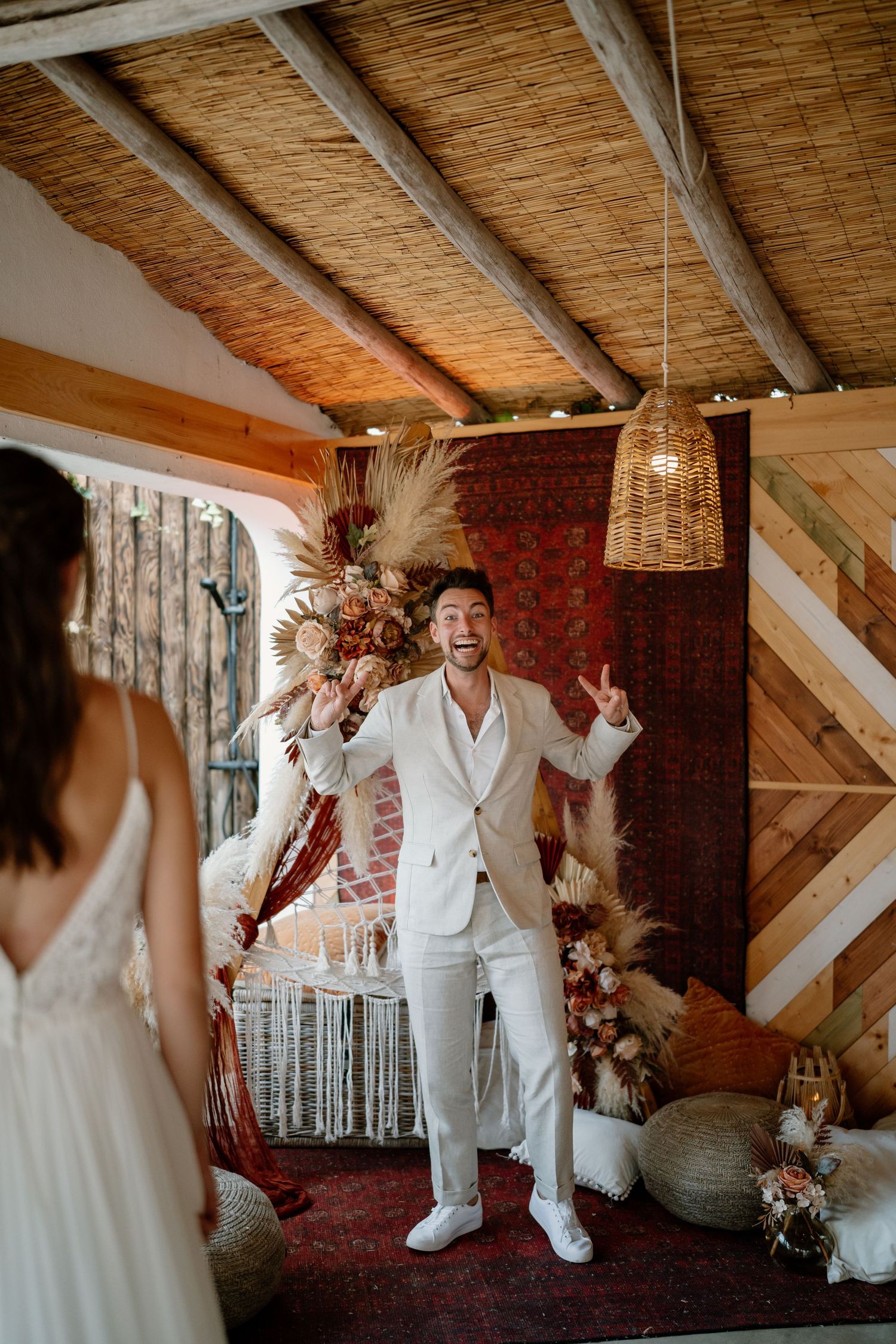 Groom in white suit reacts joyfully as bride approaches in boho wedding venue with pampas grass decor.