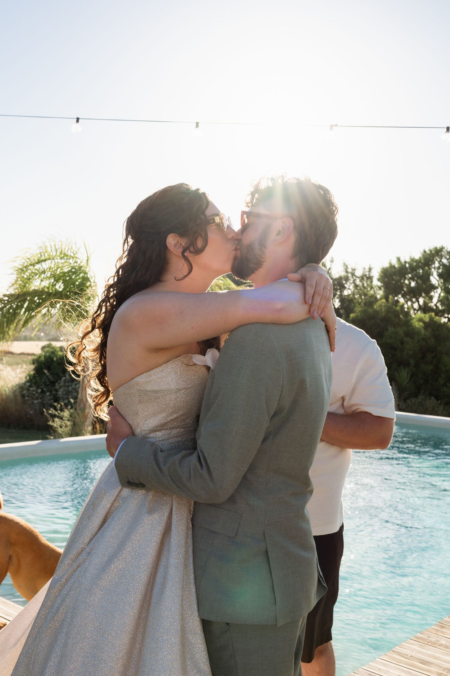 Bride and groom sharing a romantic kiss by a sunlit pool during their outdoor wedding ceremony.