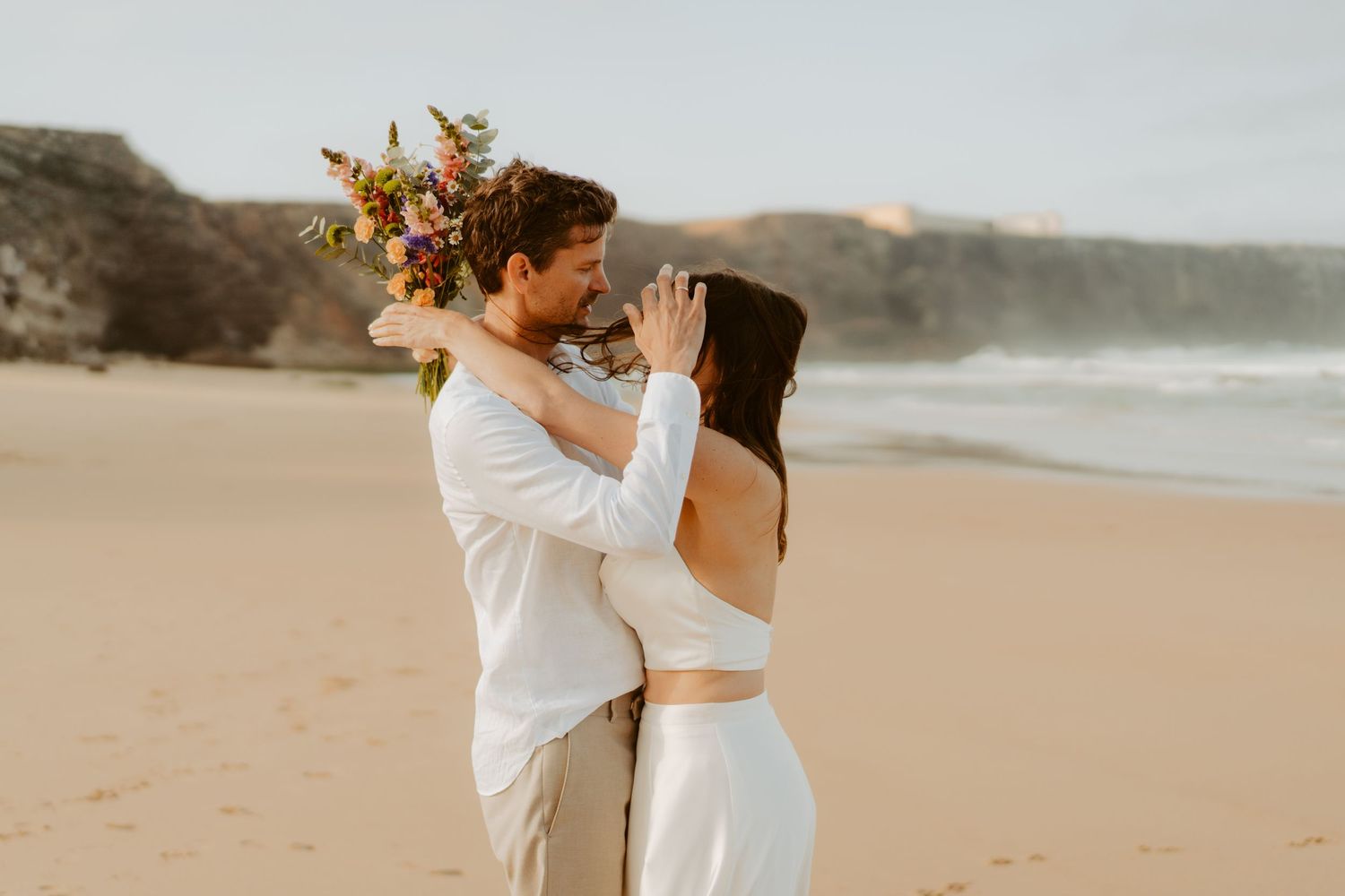 Wedding couple share a romantic embrace on a beach, bride holds a vibrant bouquet with ocean in background.