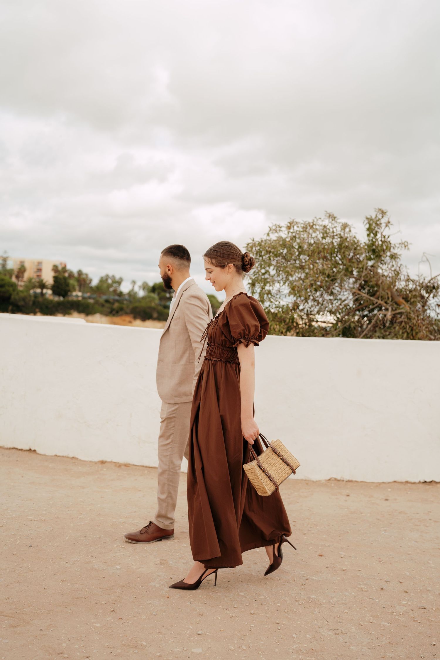 Elegant couple outdoors, woman in brown maxi dress with wicker bag, man in beige suit, walking on sandy path.
