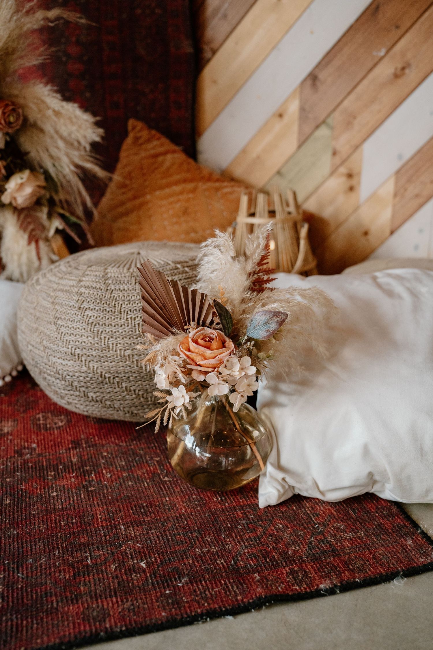 Boho dried floral arrangement in glass vase on red rug beside white pillows and wooden chevron wall.