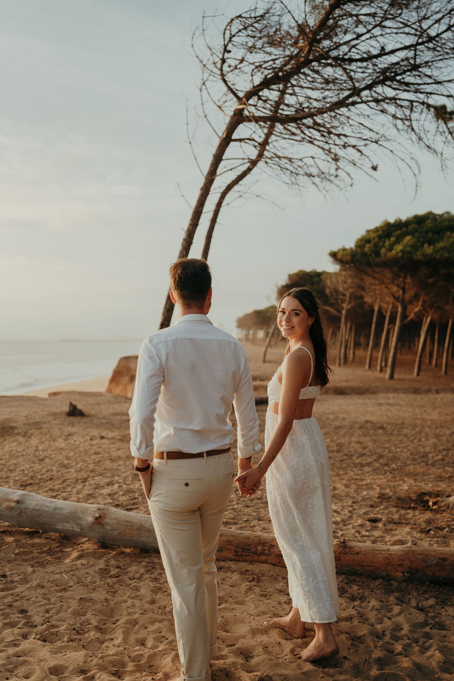 Couple holding hands on a sandy beach at sunset, woman in white dress smiling, trees in background.