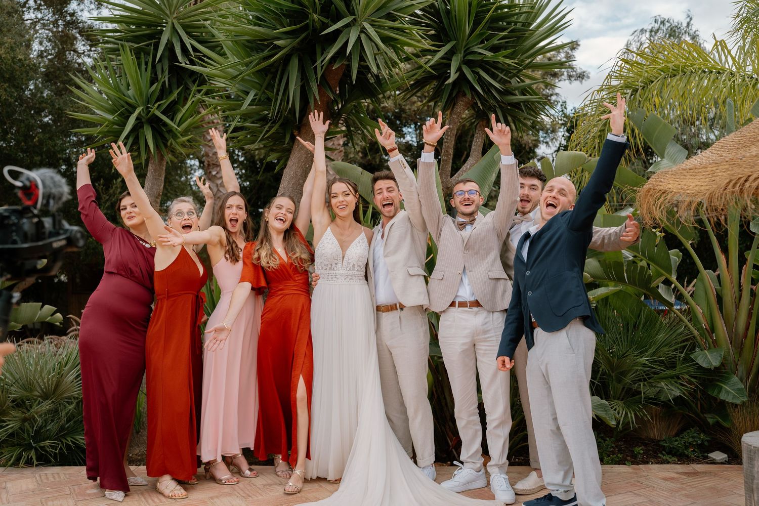 Joyful wedding party celebrating outdoors in colorful dresses and suits, raising arms in excitement among tropical greenery.