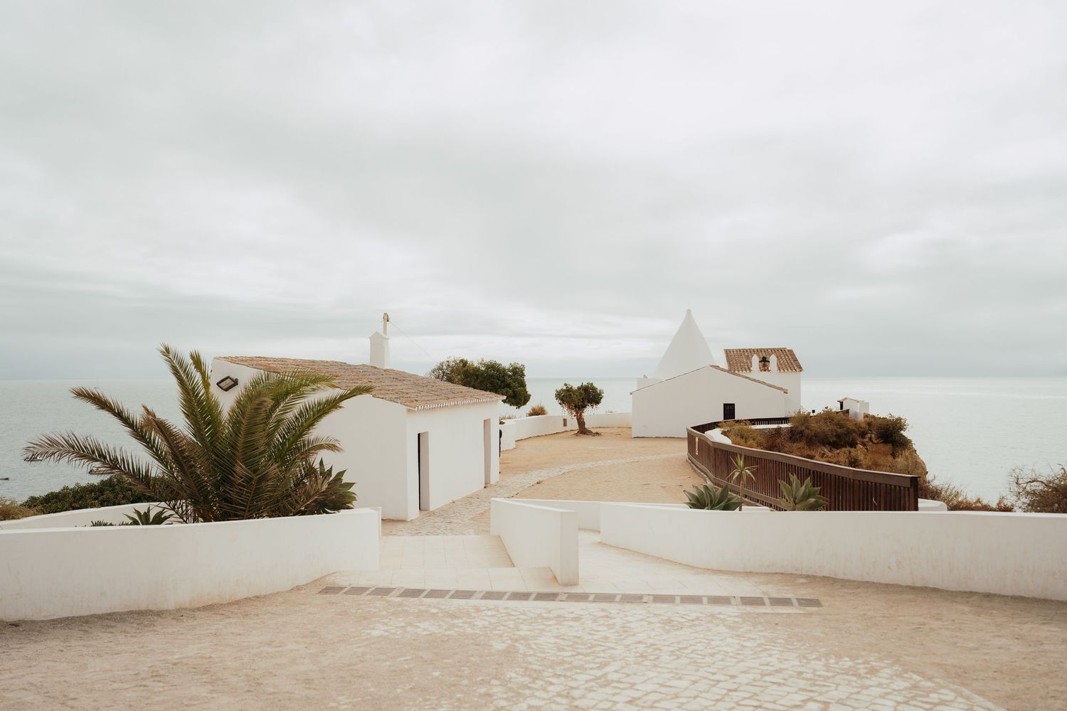 White Mediterranean buildings on a coastal hilltop with palm trees and ocean views under an overcast sky.