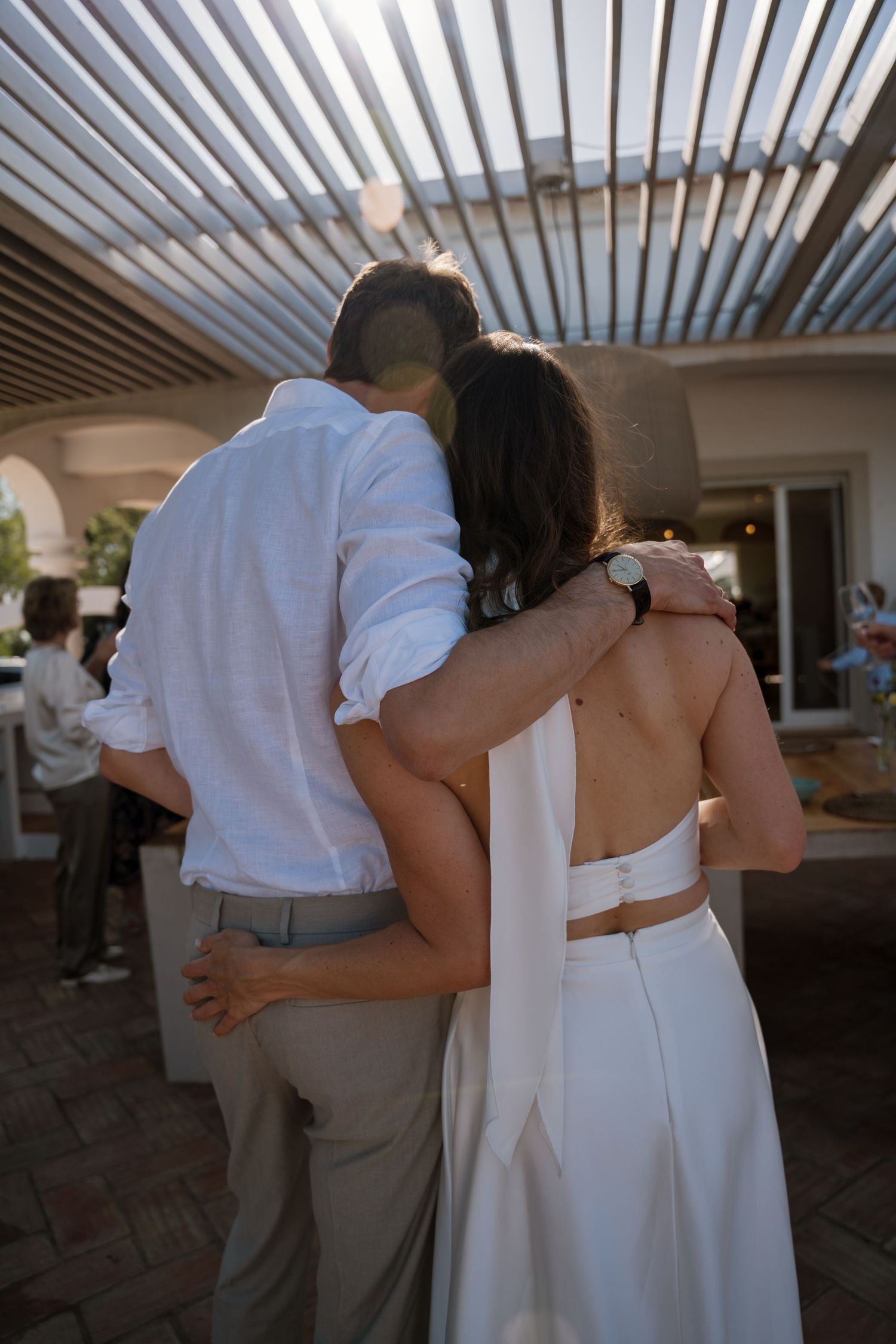 Bride and groom embrace from behind under a pergola at an outdoor wedding reception.
