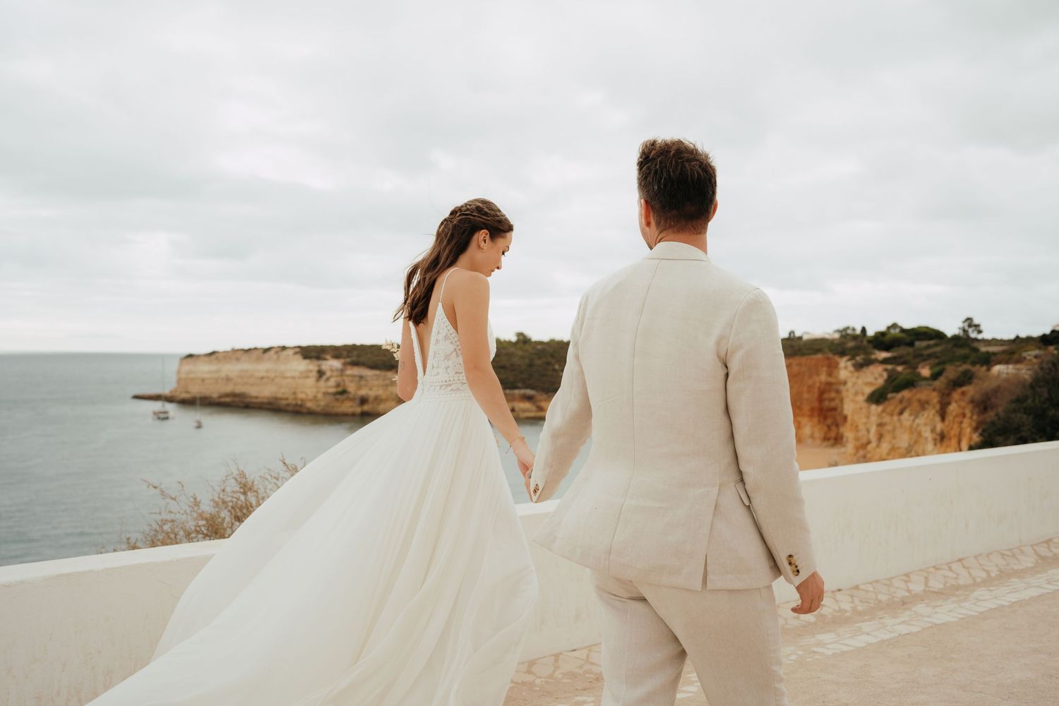 Bride in a flowing white gown and groom in a beige suit hold hands walking along a coastal terrace.