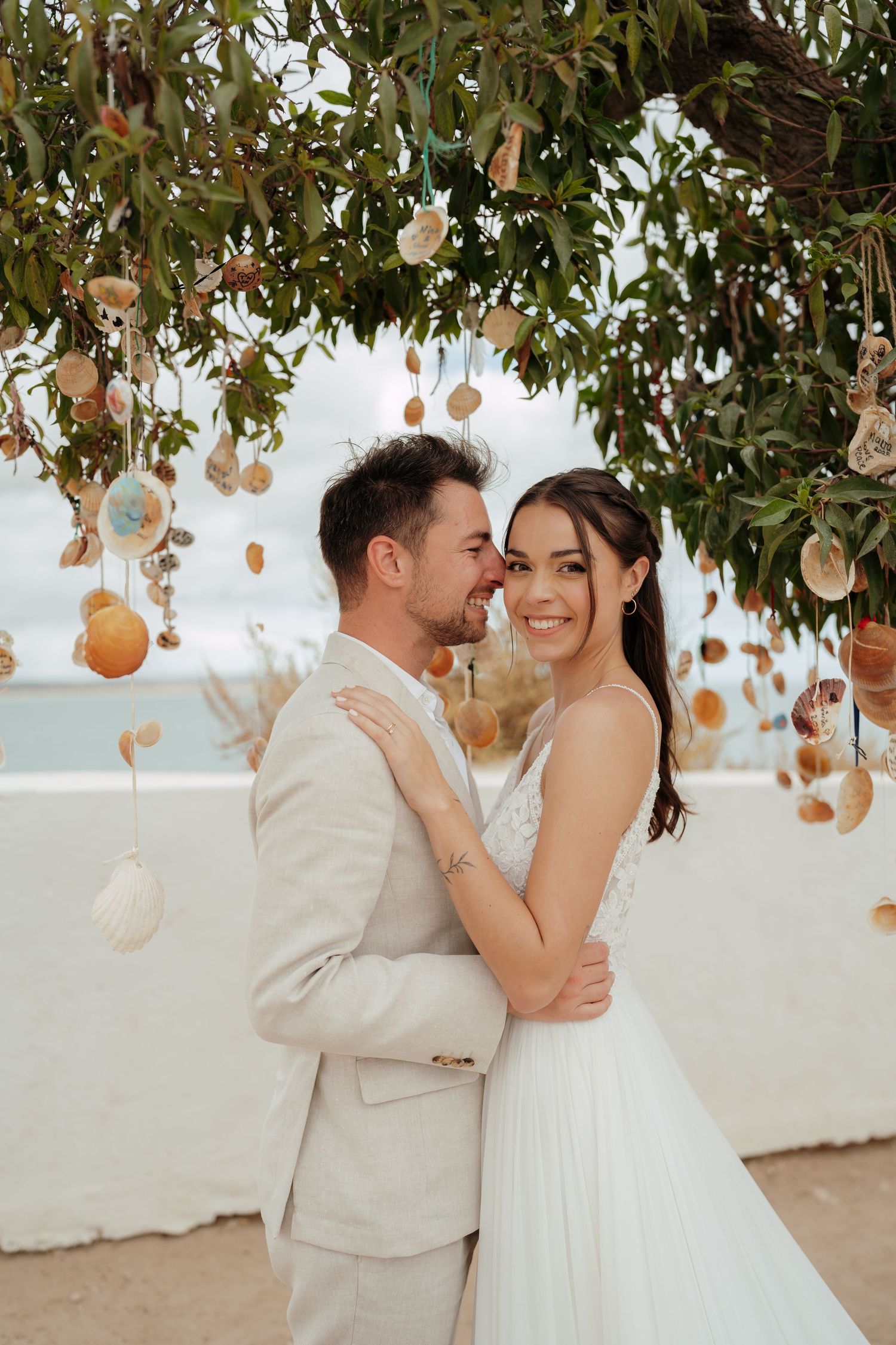 Bride and groom embrace under a tree decorated with hanging seashells at a beach wedding venue.