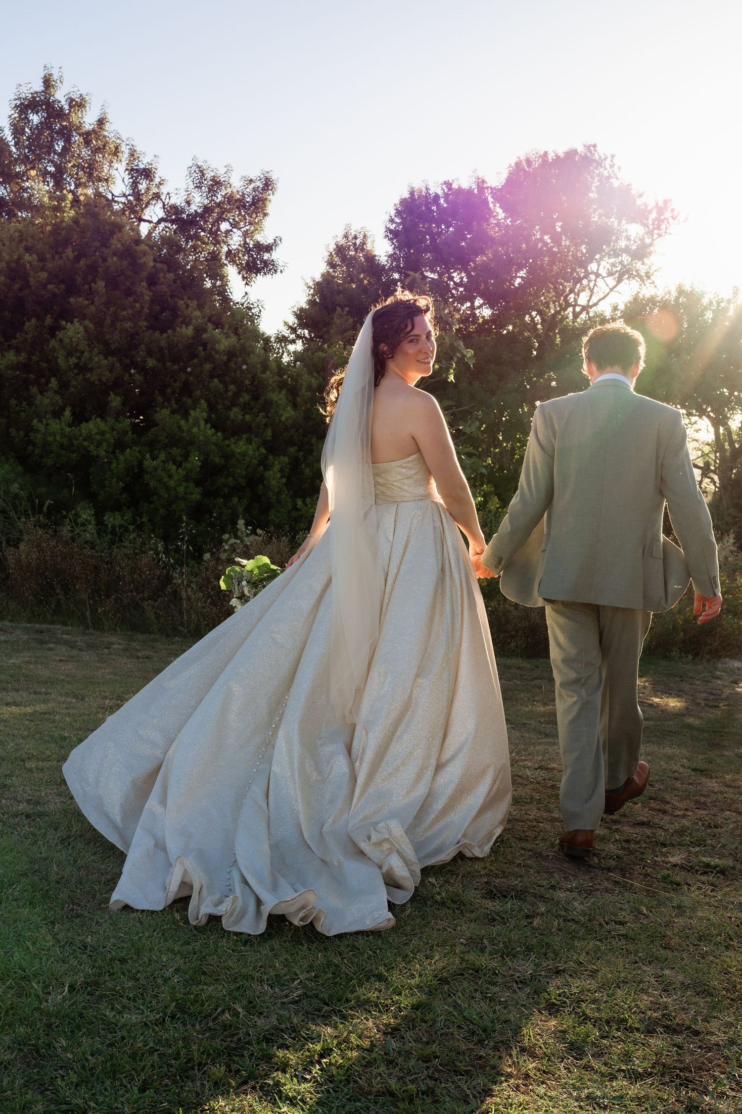 Bride and groom walking hand in hand through lush garden at sunset, bride in flowing white gown with veil.