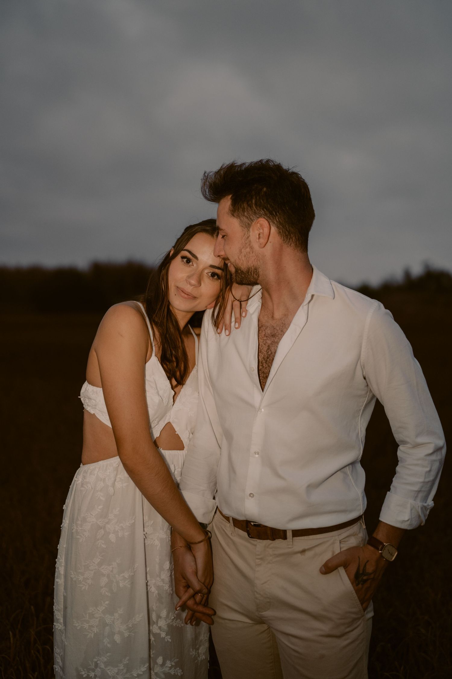 A couple in white attire embraces in a field at dusk, man kissing womans temple while holding hands.