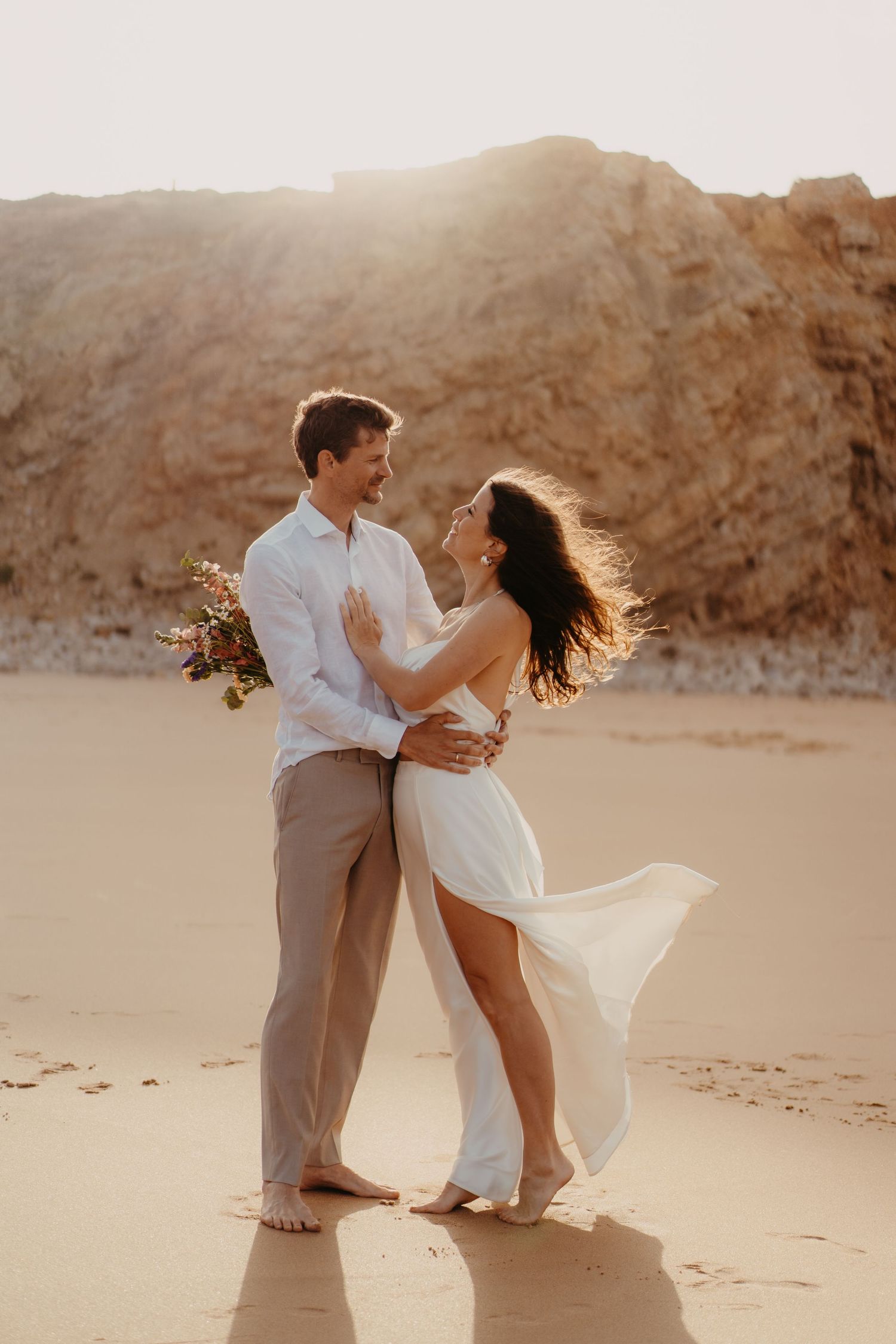 Couple embracing on sandy beach at golden hour, bride in white flowing dress holding bouquet.