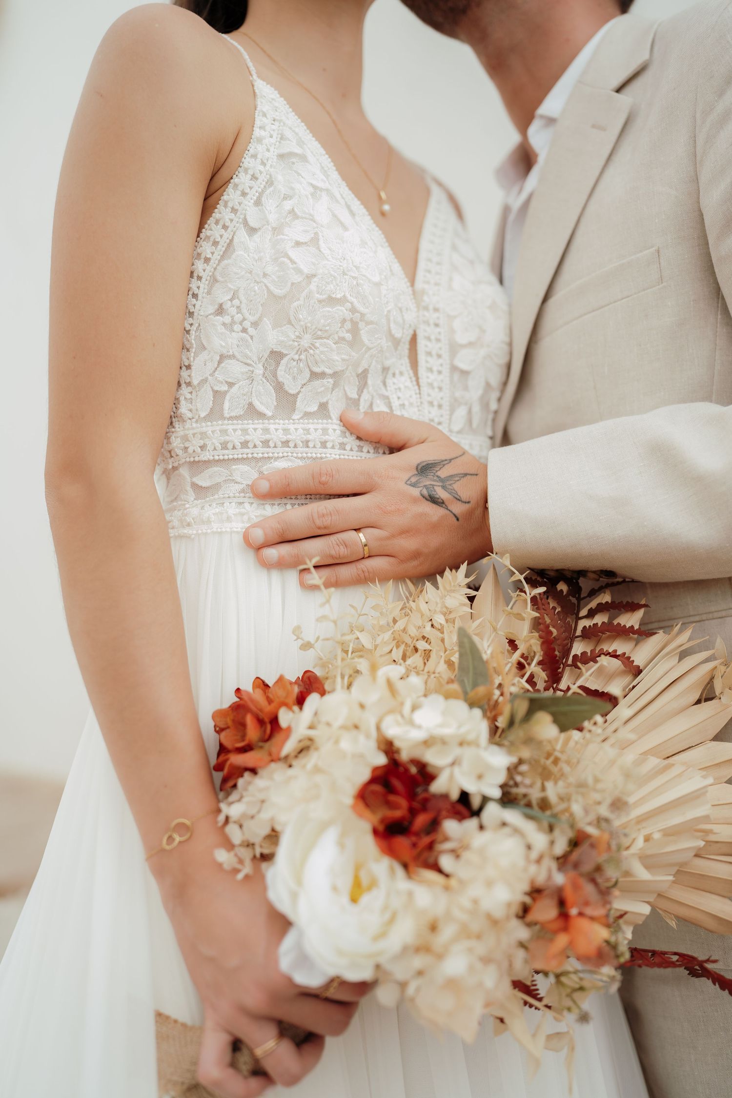 Bride and groom embracing, holding a boho bouquet of white and rust flowers, bride wearing lace wedding dress with ring visible.