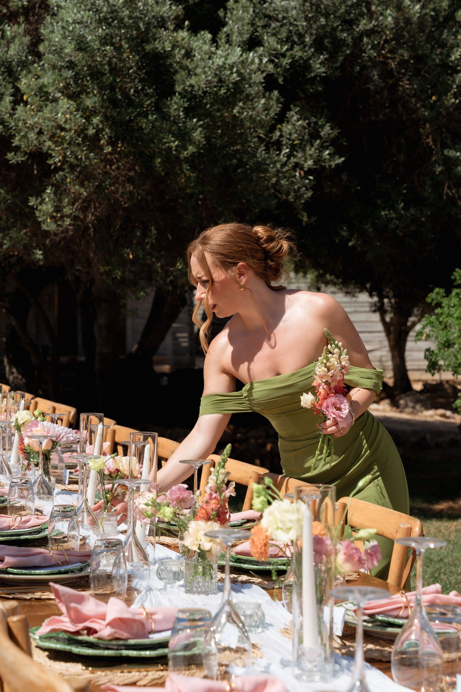 Woman in green off-shoulder dress arranging floral centerpieces on an elegant outdoor wedding reception table.