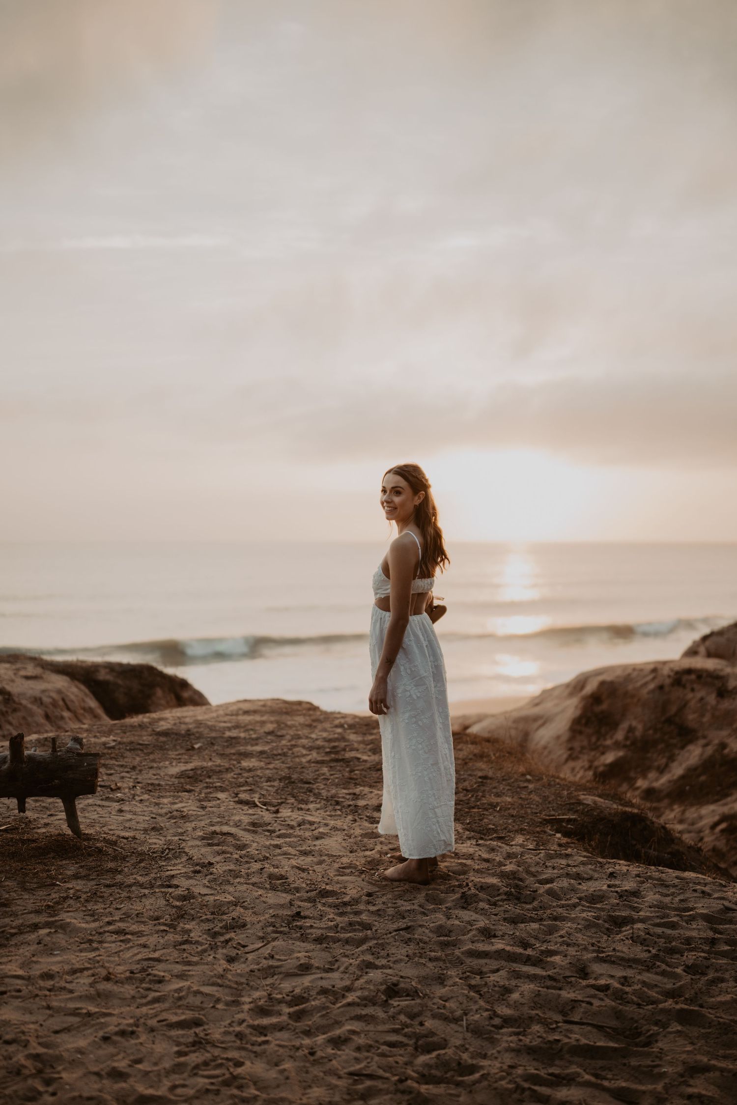 Woman in white dress standing on sandy beach rocks at golden sunset with ocean waves in background.