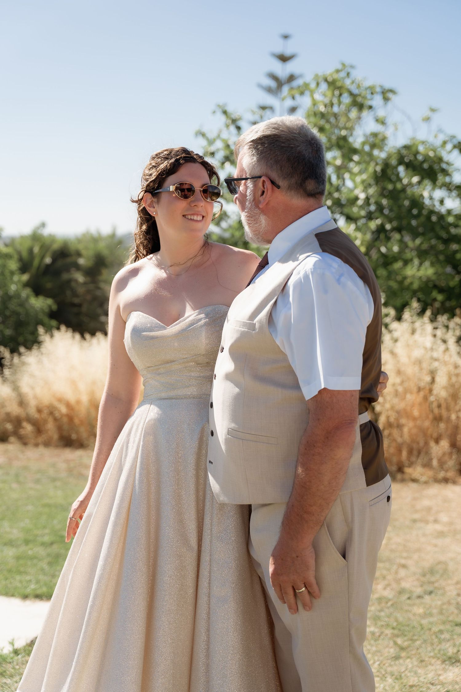 Bride and groom in sunglasses share a romantic moment outdoors on a sunny wedding day.