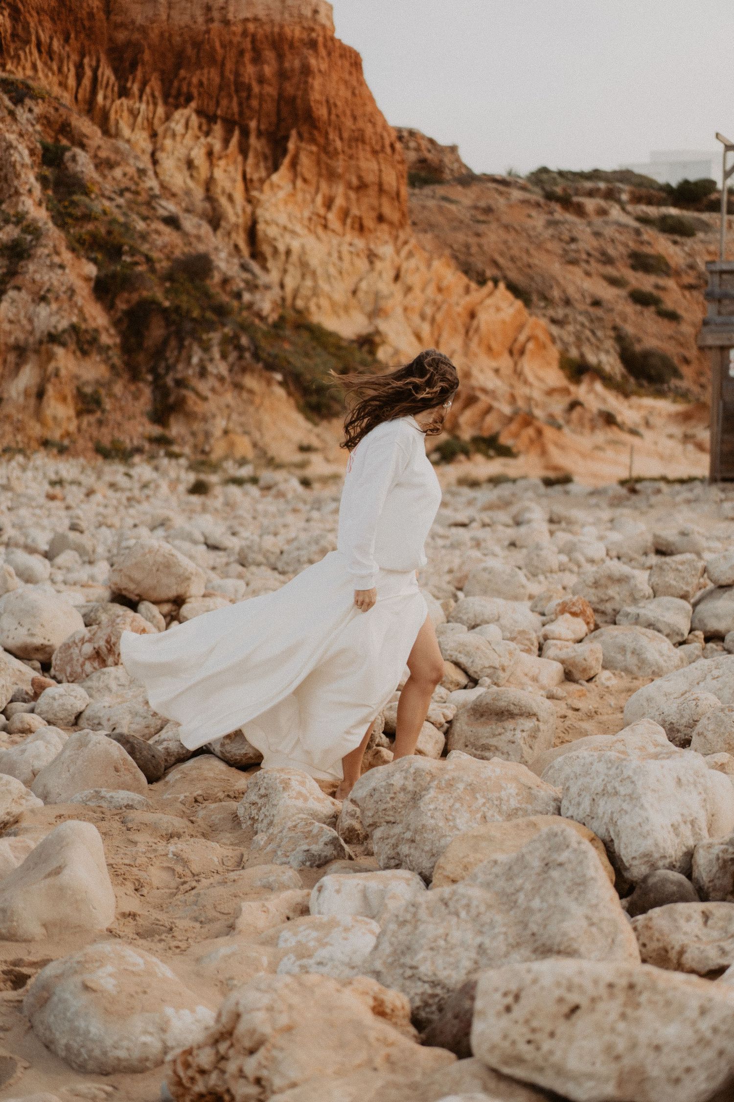 Bride in flowing white dress walking on rocky beach with dramatic red cliffs in the background.