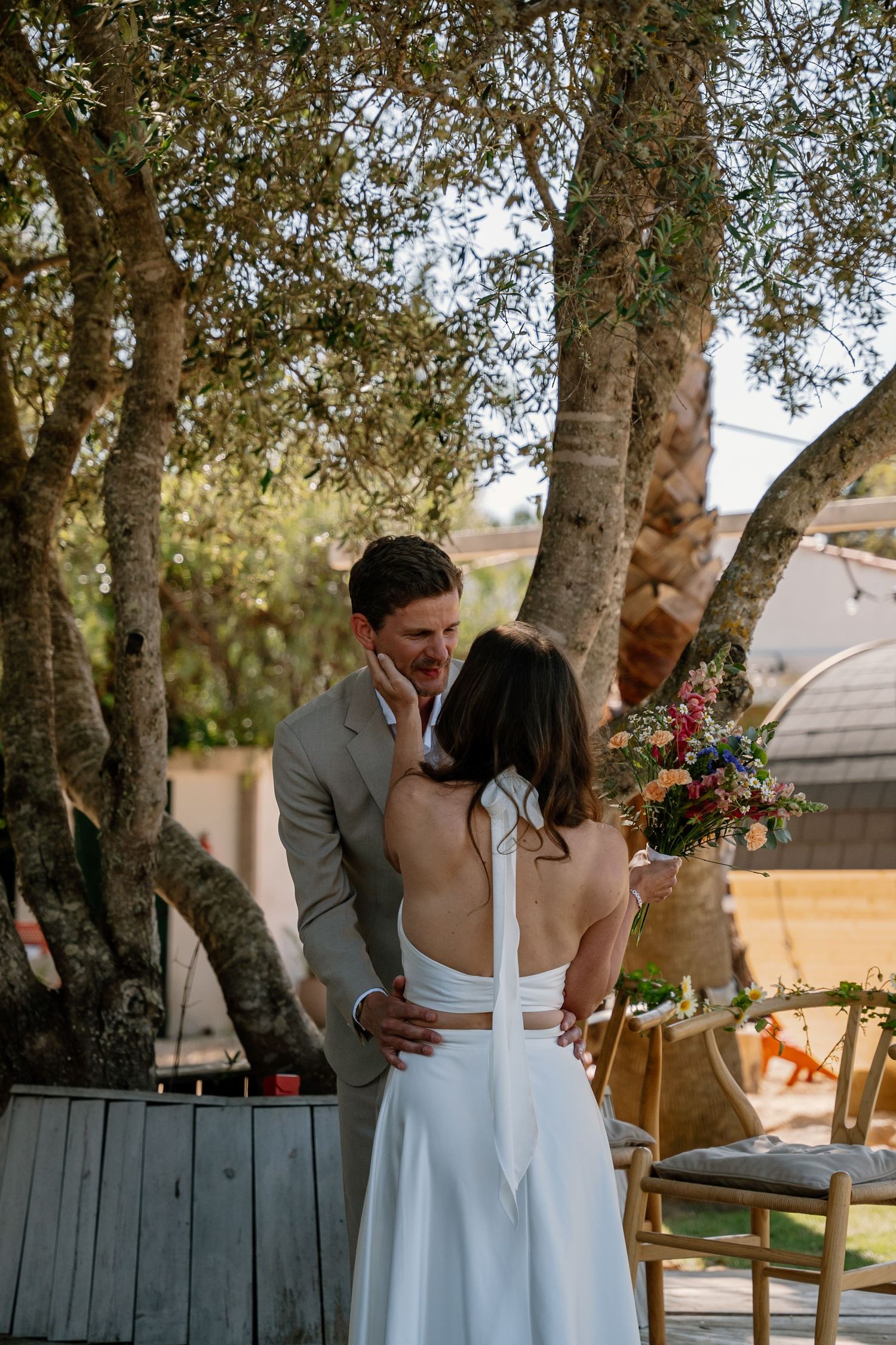 Groom in tan suit tenderly touches brides face during intimate outdoor wedding ceremony under olive trees.