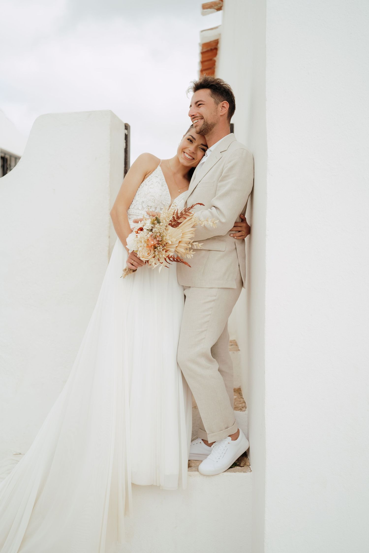 A bride and groom pose together on white steps, she holds a dried floral bouquet in a flowing white gown.
