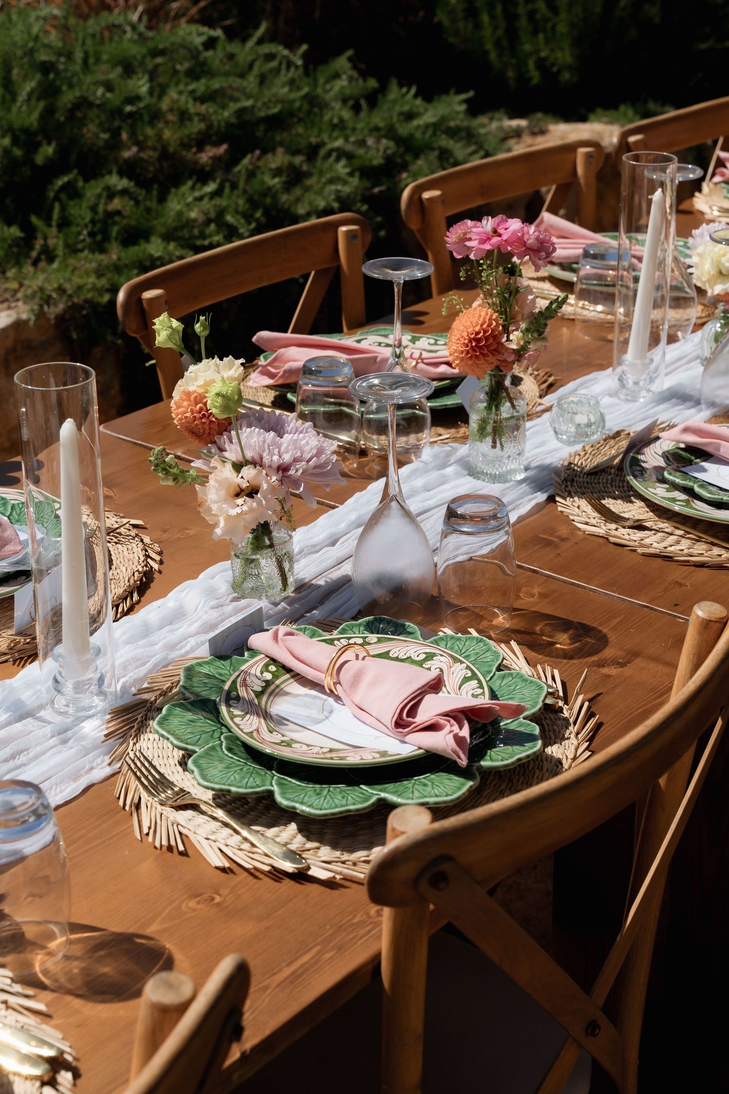 Elegant outdoor dining table set with green plates, pink napkins, woven placemats, and fresh floral centerpieces.