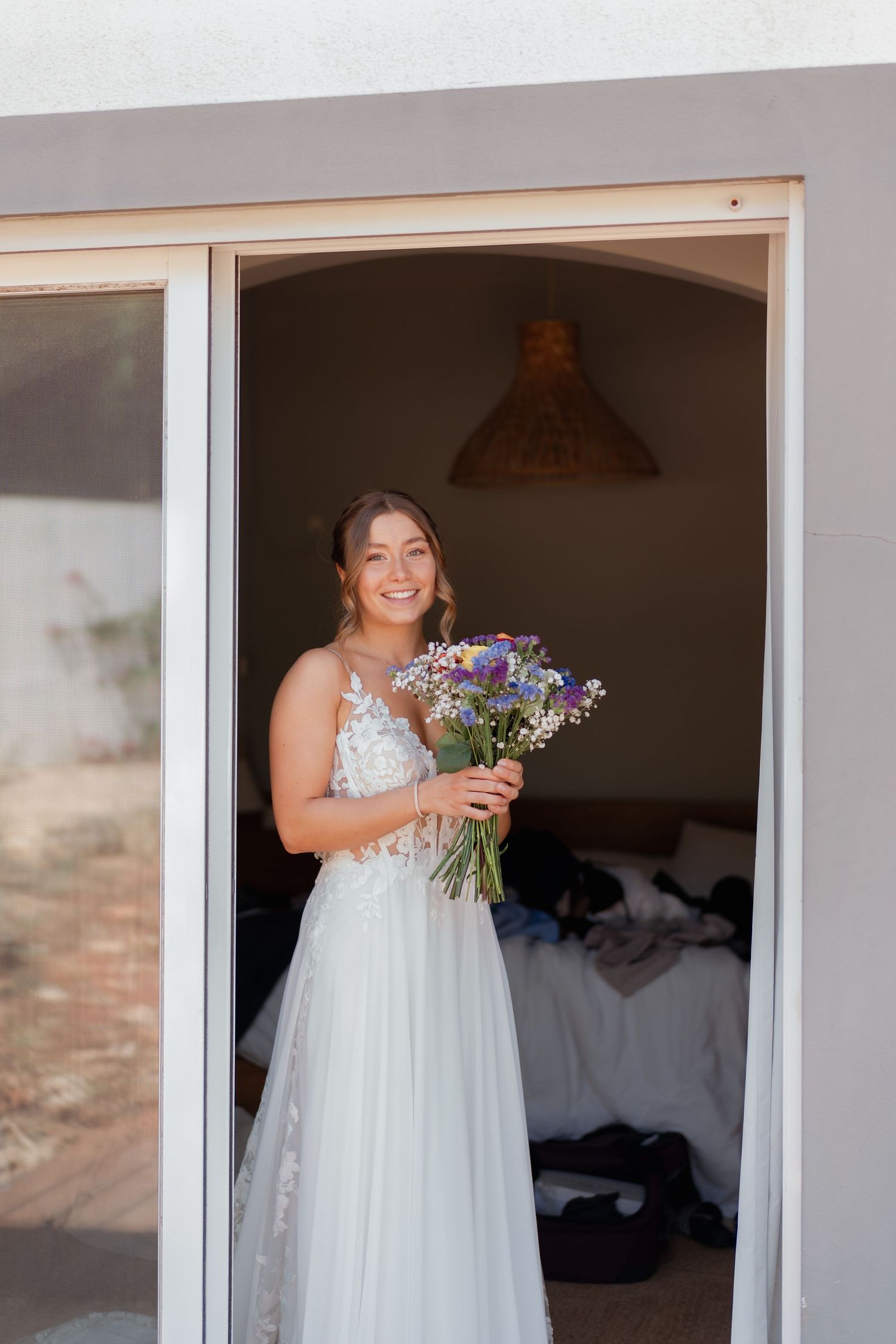Smiling bride in white lace gown holds wildflower bouquet standing in doorway of sunlit room.