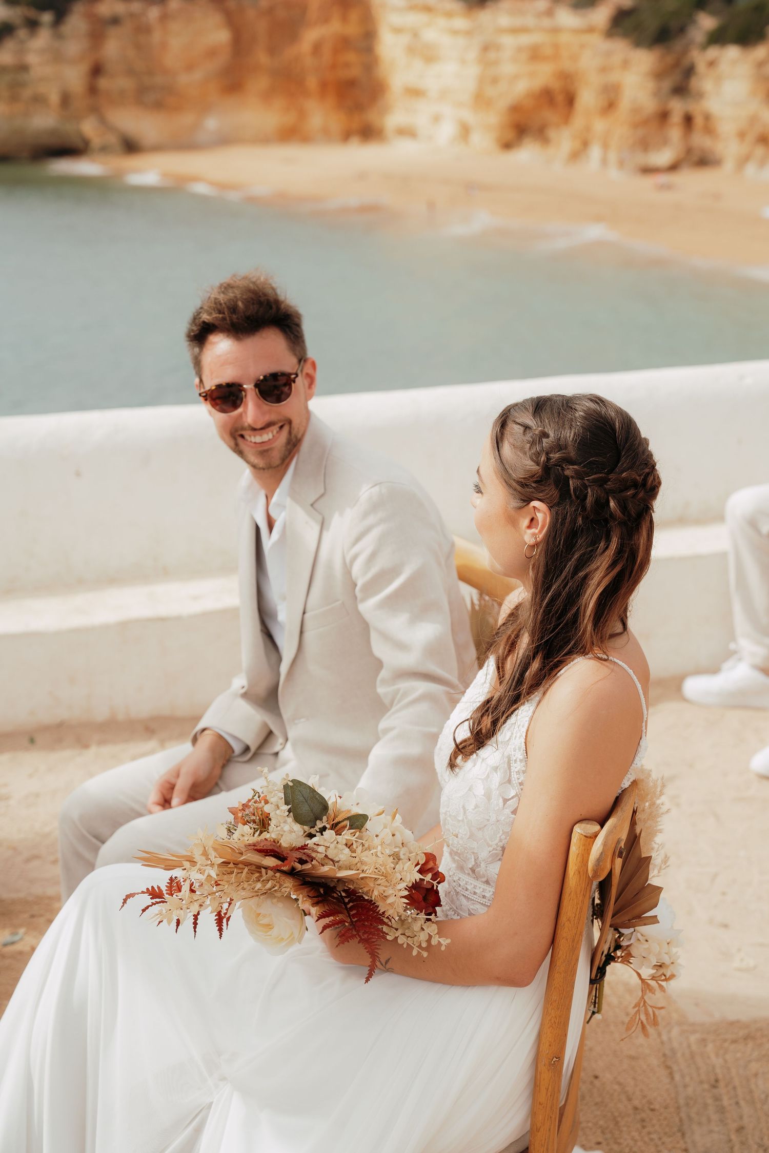 Bride and groom seated outdoors near a coastal cliff, bride holding dried floral bouquet in white dress.