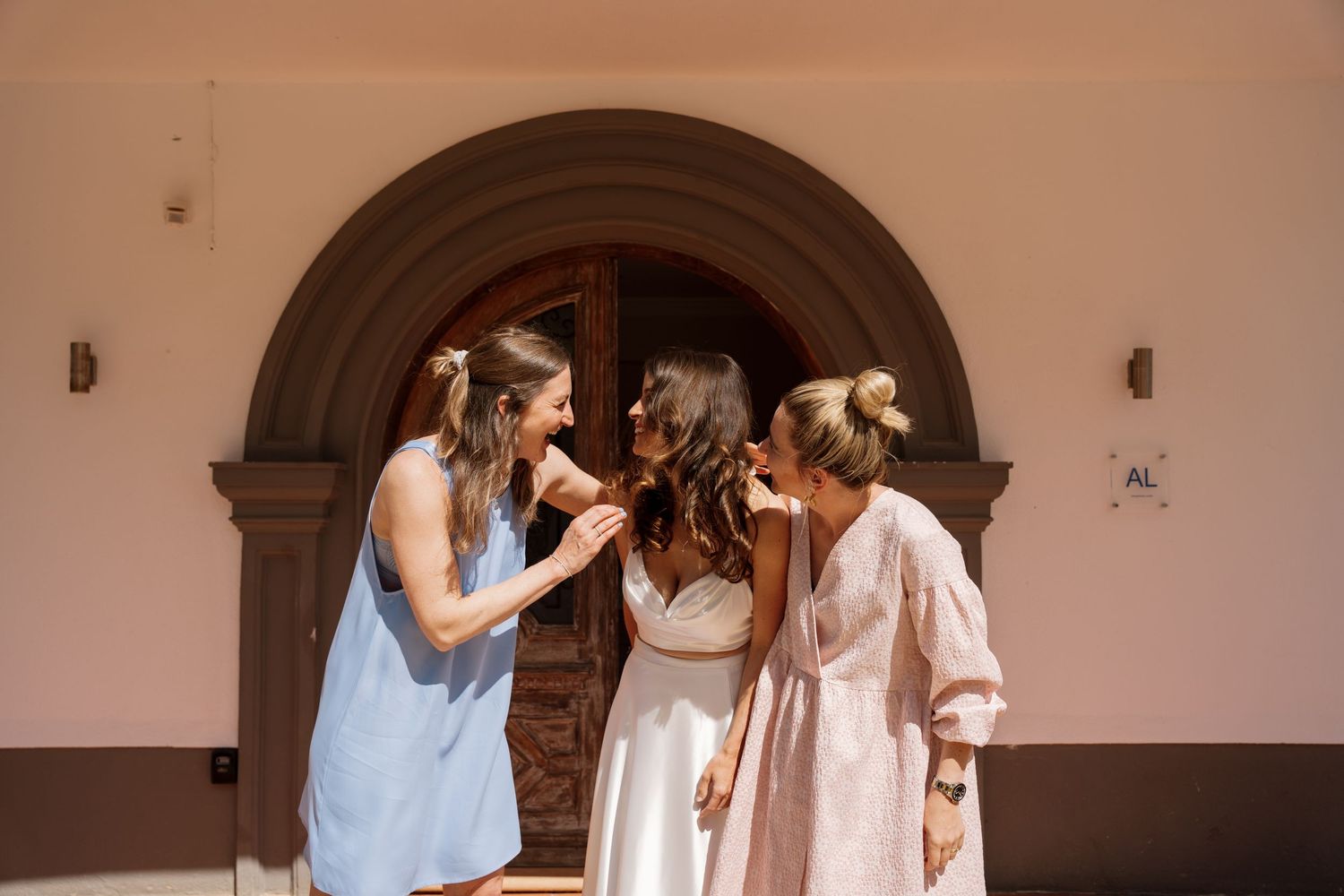 Three women laughing together outside an arched doorway on a sunny day.