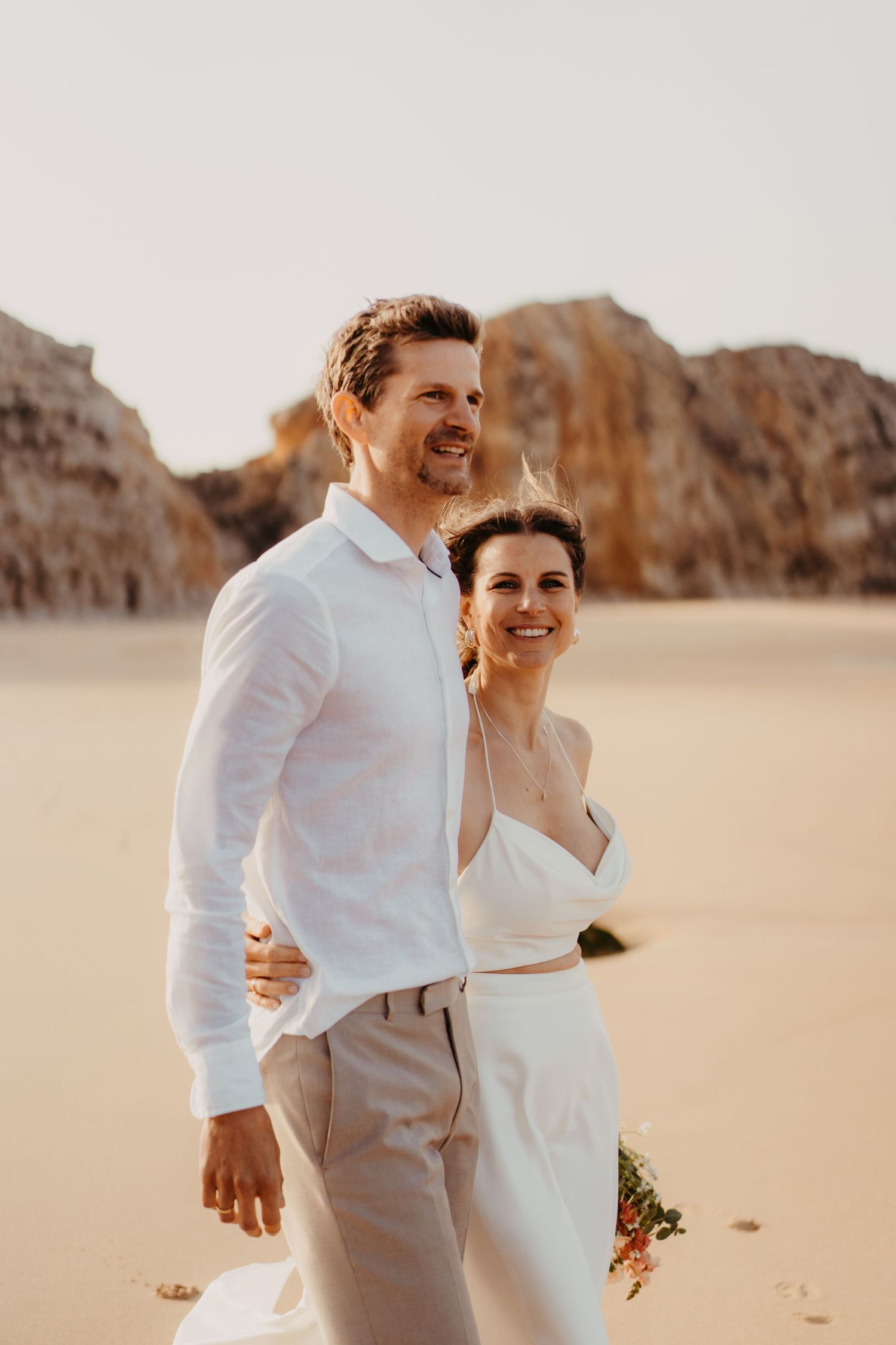 Happy couple in wedding attire embrace on a sandy beach with rocky cliffs glowing in warm sunset light.