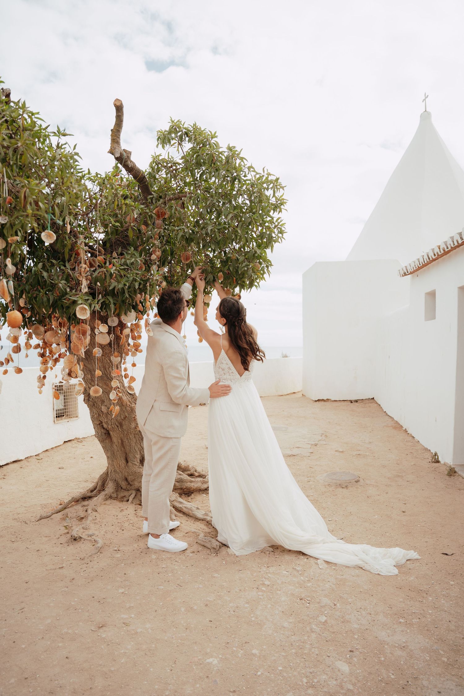 Bride and groom stand beneath a decorated tree with hanging ornaments at a whitewashed outdoor wedding venue.