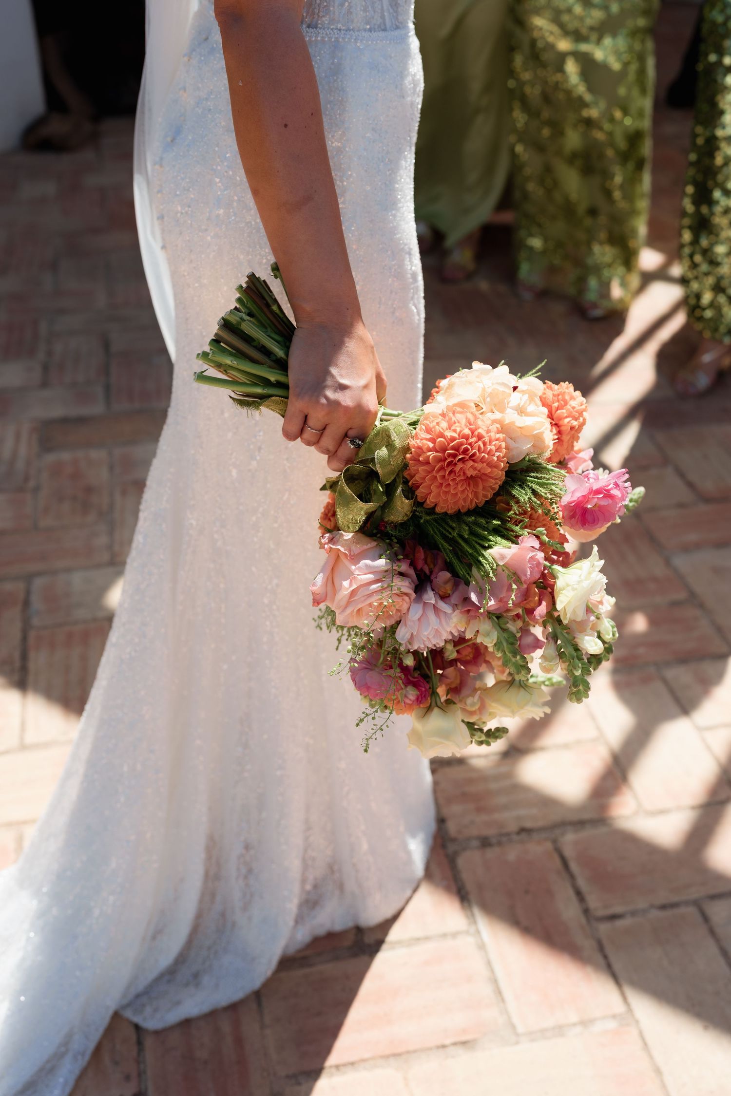Bride in white sequin gown holding a vibrant bridal bouquet with peach dahlias and pink roses on brick floor.