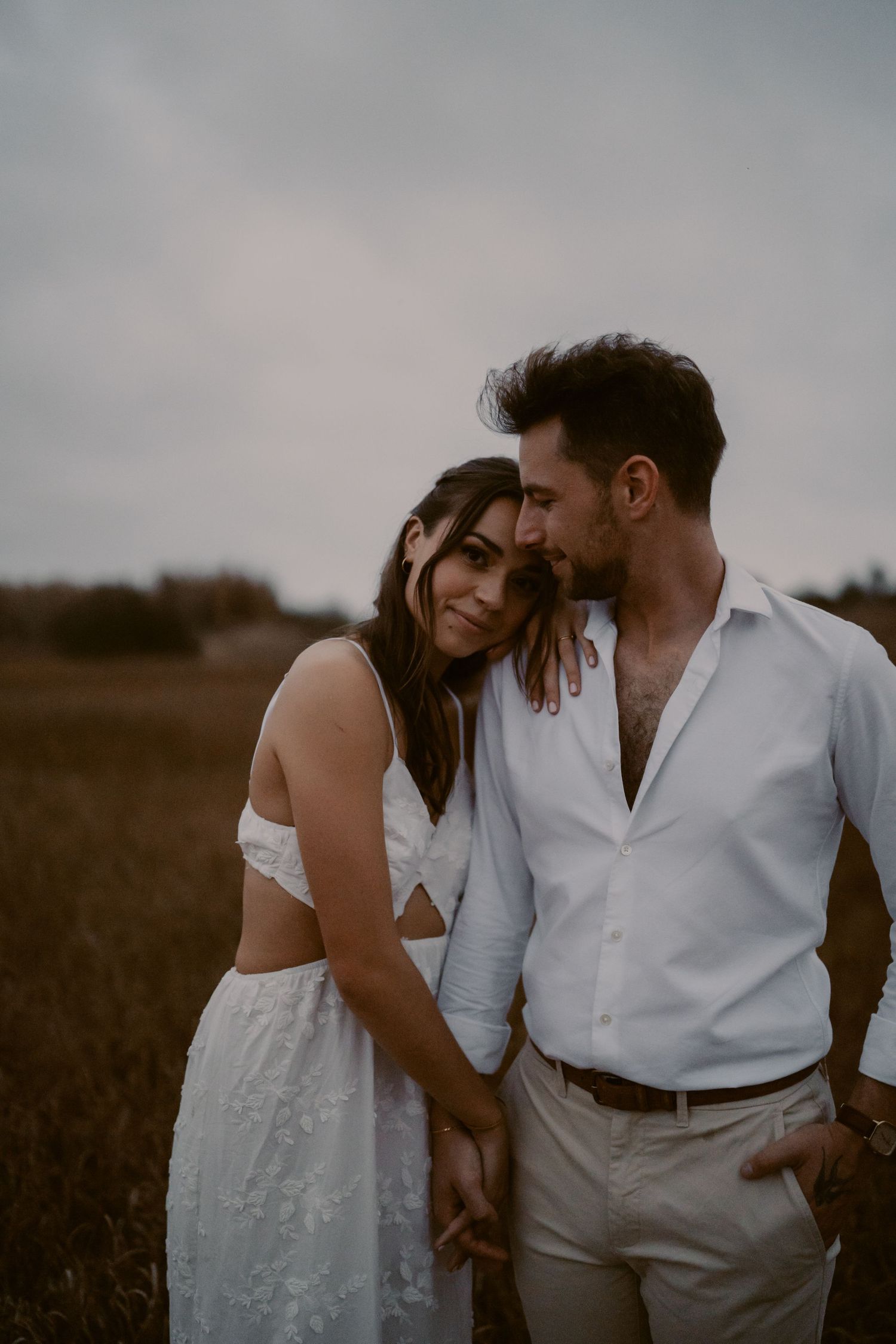 Romantic couple in white outfits embracing in a field, woman smiling softly as man leans in close on overcast day.