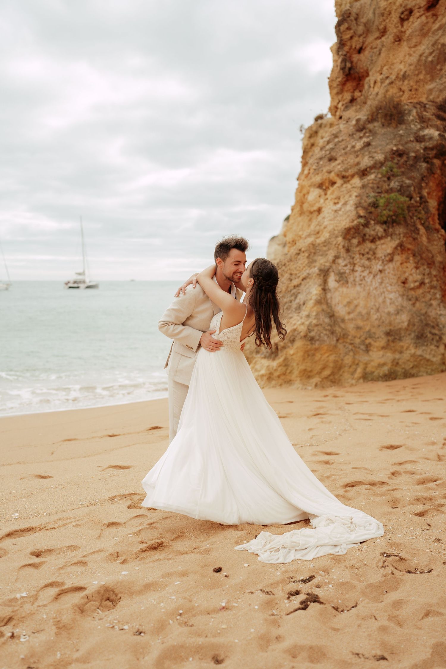 Bride and groom share a romantic kiss on a sandy beach with ocean waves and rocky cliffs in the background.