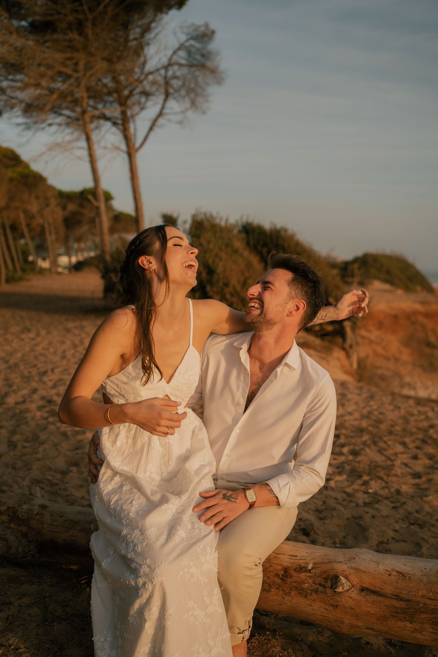 A joyful couple in white outfits laughing together on a sandy beach at golden hour sunset.