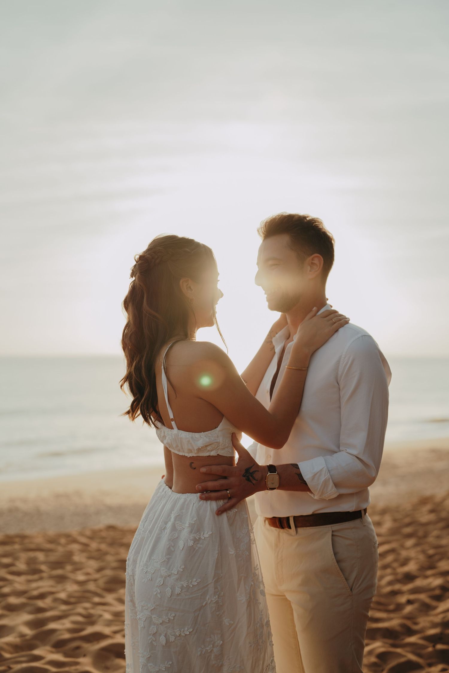 Couple in wedding attire embracing on a sandy beach at golden sunset, woman in white lace dress facing man in light shirt.