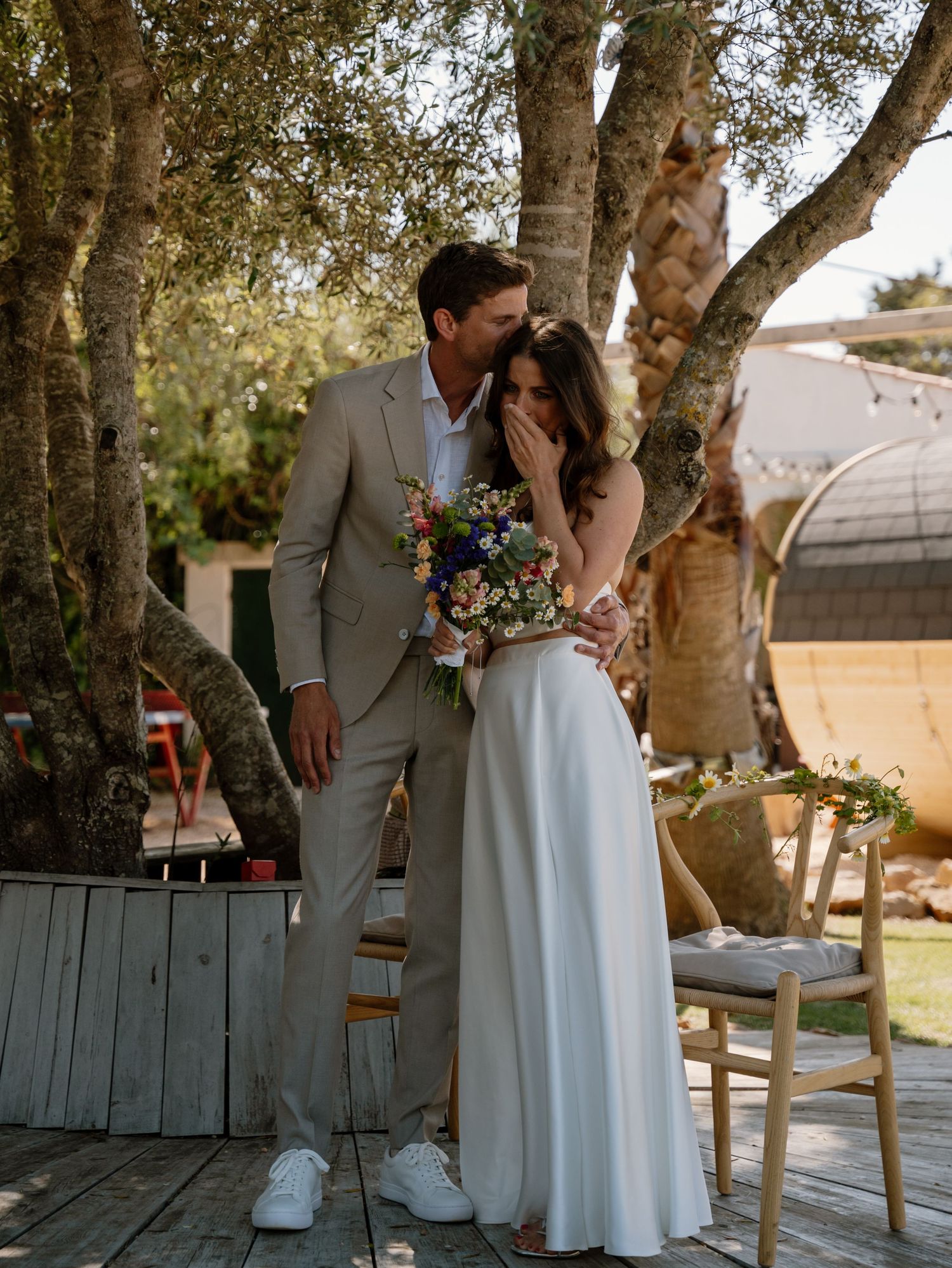 Groom embraces emotional bride holding a wildflower bouquet during outdoor wedding ceremony under olive trees.