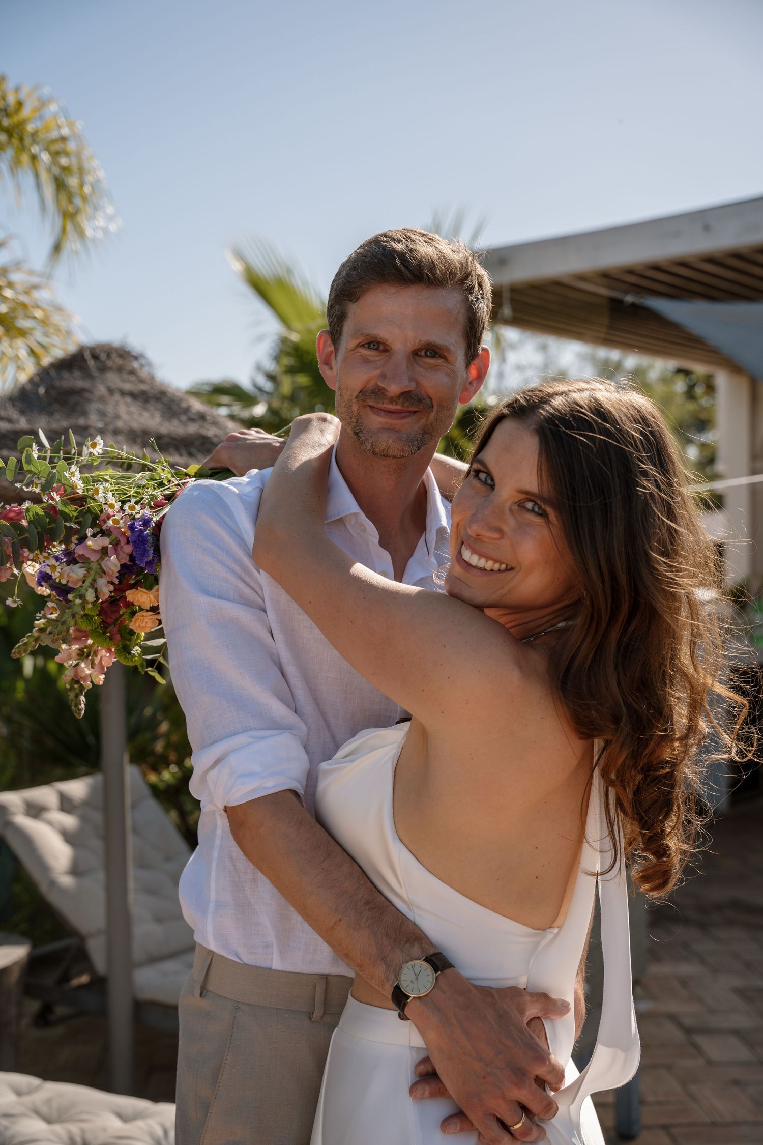 Smiling bride and groom embrace outdoors holding a colorful wildflower bouquet at a beach wedding.