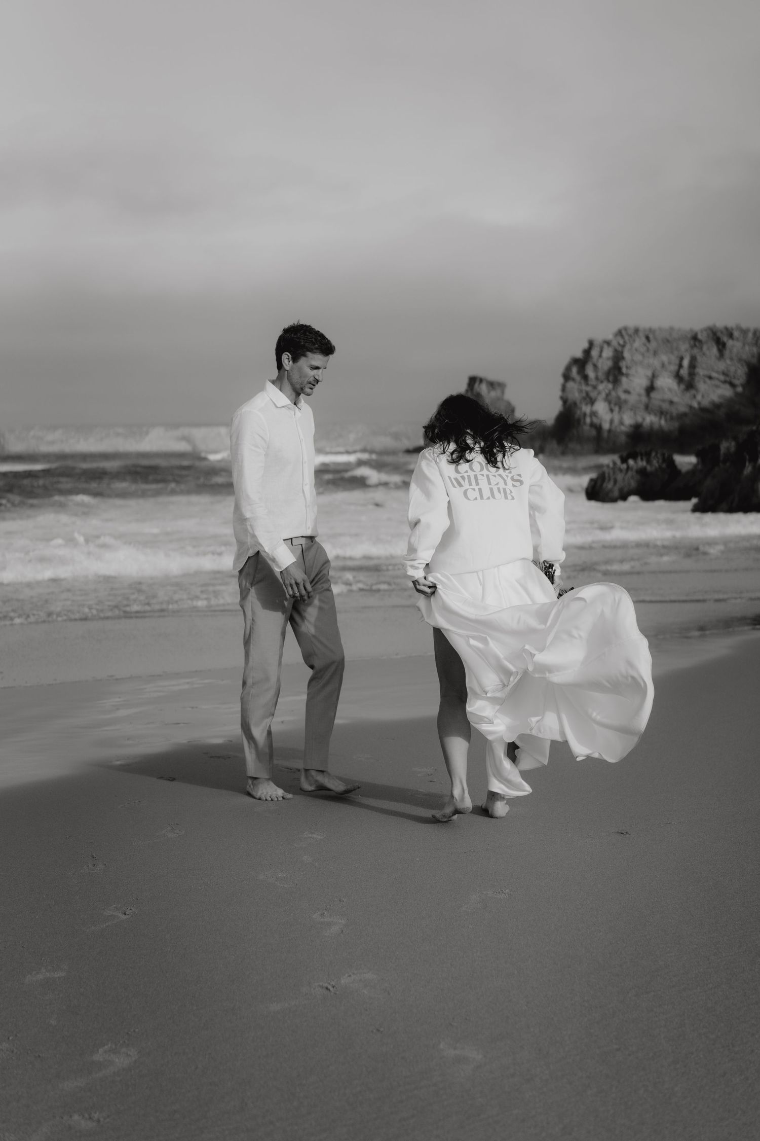 Black and white photo of bride in wifes jacket dancing on beach while groom watches and smiles.