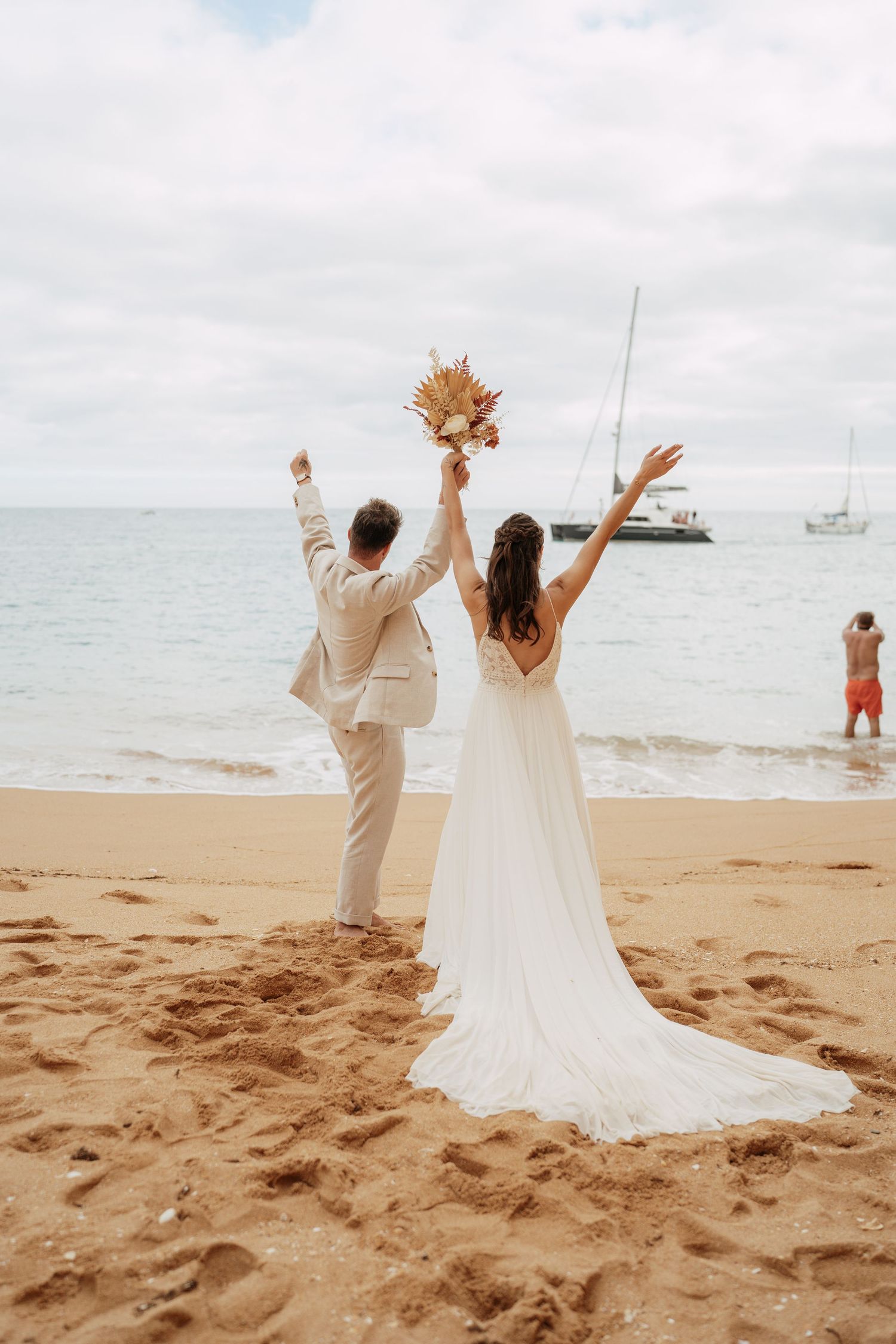 Bride and groom celebrate on sandy beach, arms raised, holding bouquet, with ocean and sailboat backdrop.