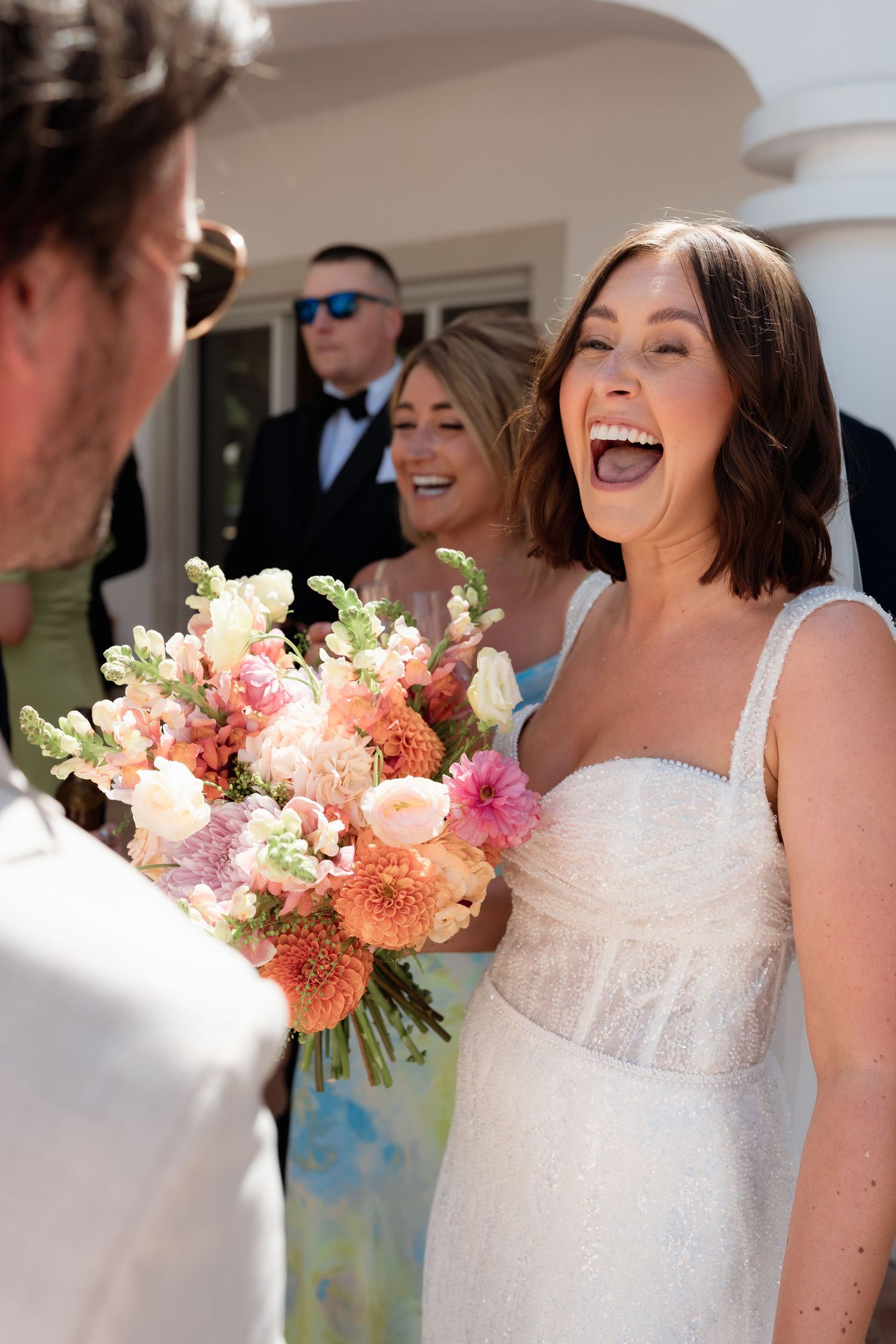 Joyful bride in white gown laughing while holding colorful pink bouquet with bridesmaids at sunny outdoor wedding.