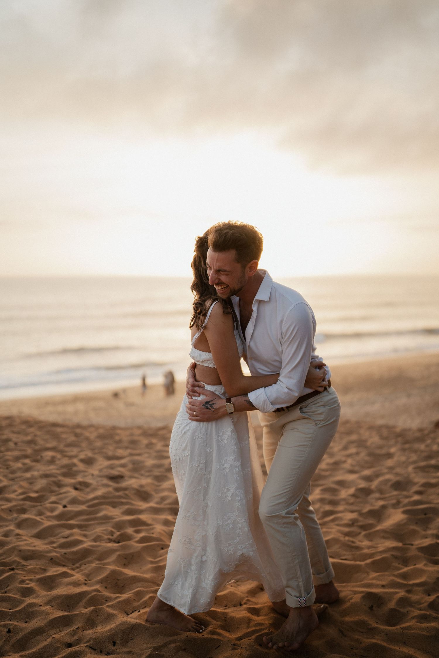 Couple in wedding attire embracing on sandy beach at golden sunset, waves and soft sky in background.