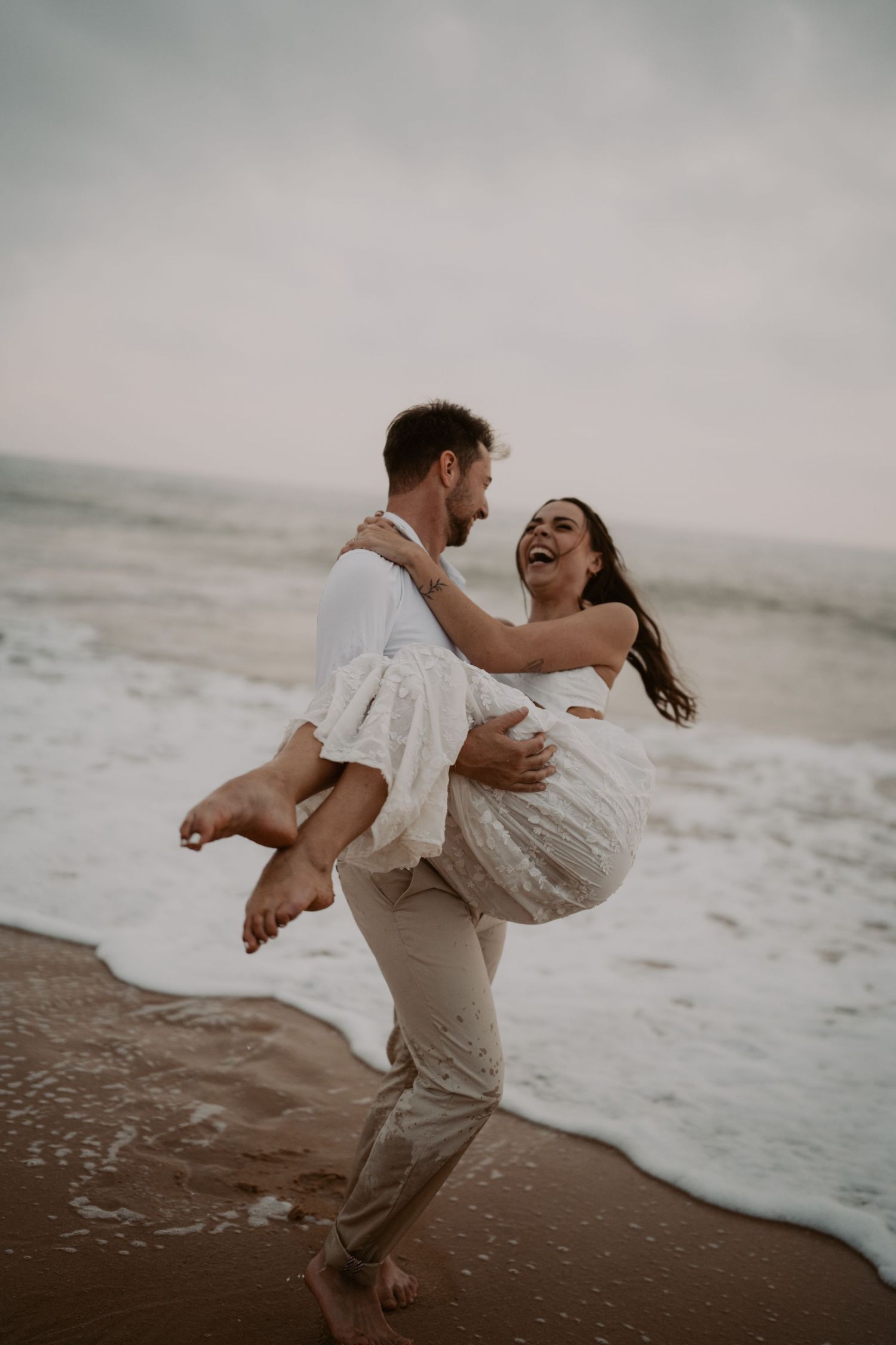 A joyful couple in white wedding attire laughing on a beach, the groom carrying the bride near ocean waves.
