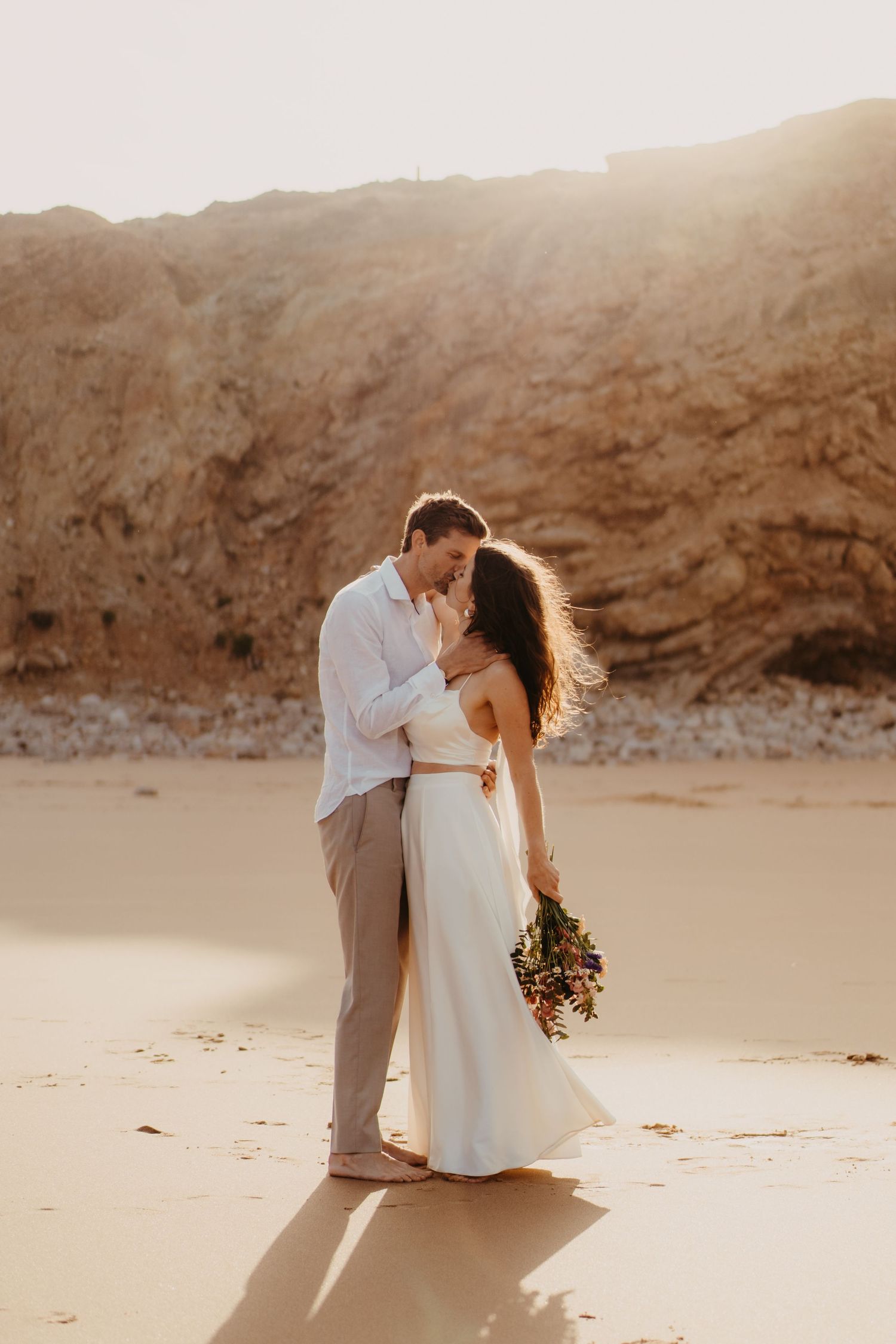 Bride and groom embrace on sandy beach at golden hour, she holds a bouquet with rocky cliffs behind them.