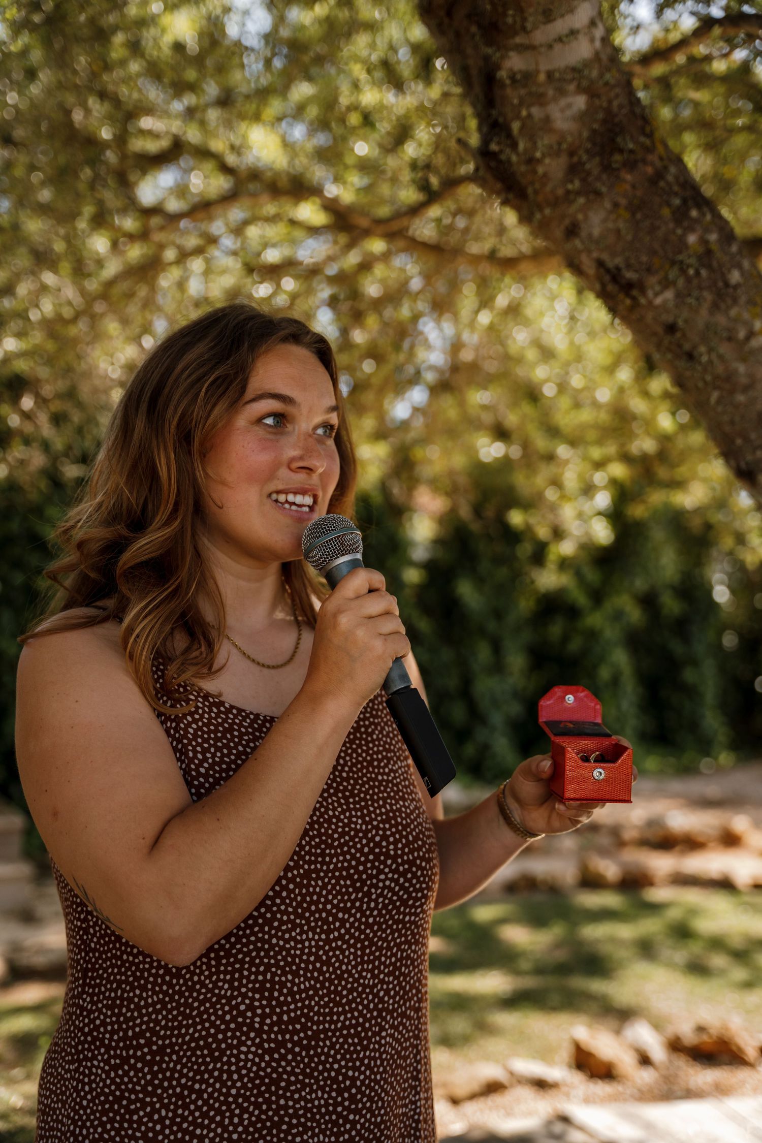 Woman in floral dress holds a red ring box and microphone during an outdoor wedding ceremony.