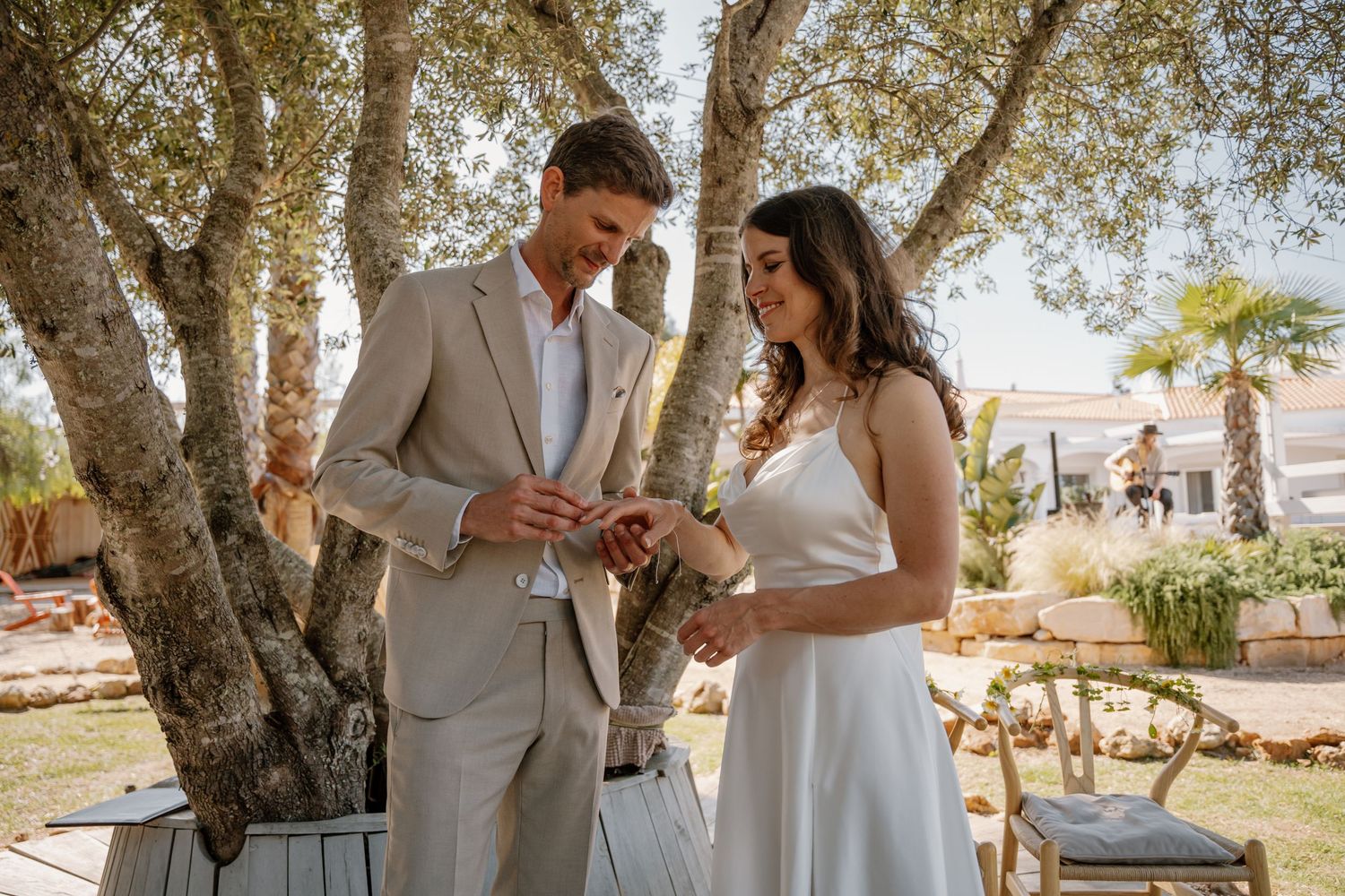 Groom places a wedding ring on the brides finger beneath olive trees at a sunny outdoor ceremony.