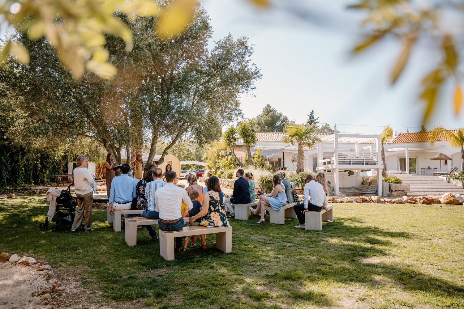 Guests seated on wooden benches in a sunny garden during an outdoor wedding ceremony at a white villa.
