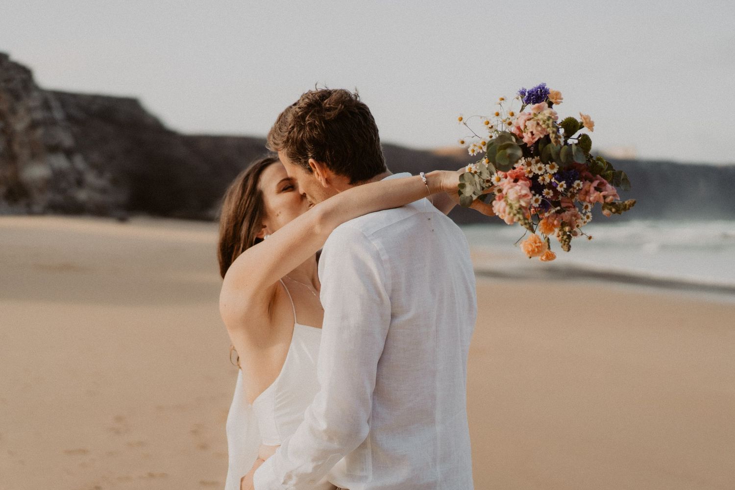 Bride and groom share a tender kiss on the beach, she holds a colorful wildflower bouquet.