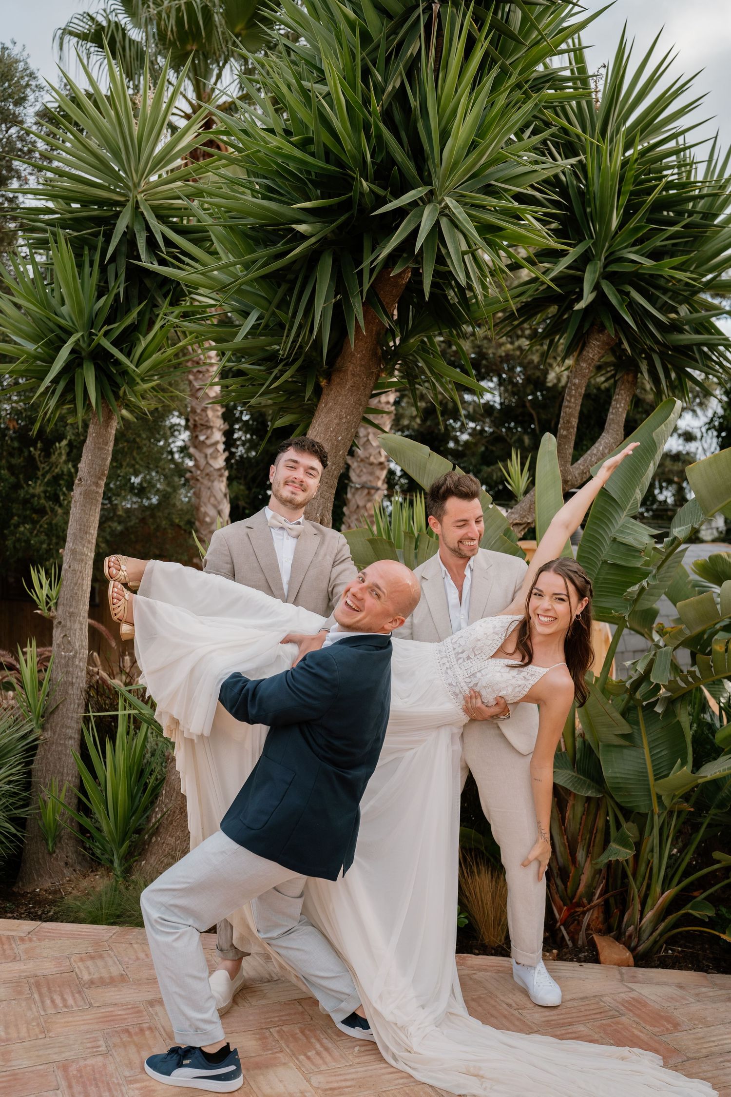 A joyful wedding group poses playfully with tropical palms, featuring bride in white gown, groom, and two guests outdoors.